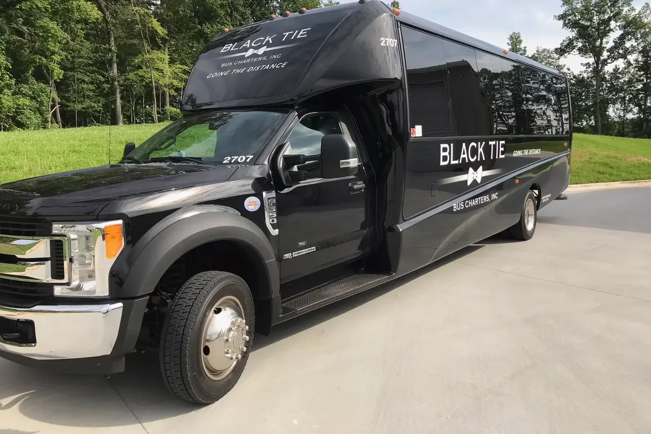 A black bus with the words 'Black Tie' and 'Bus Charters, Inc Going the Distance' written on it. The bus is parked on a paved area next to green grass and trees.