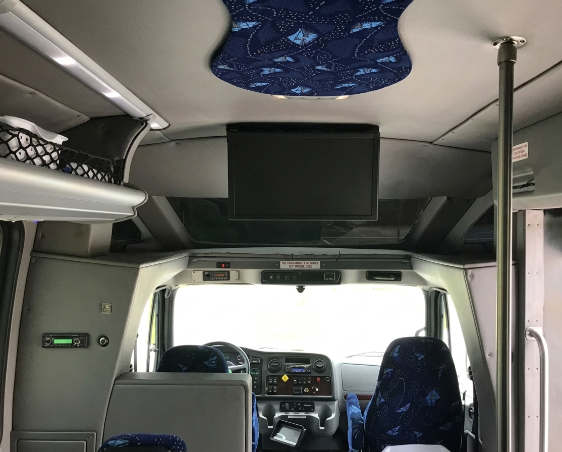 Interior of a shuttle bus with a dashboard, steering wheel, a ceiling-mounted monitor, blue and black patterned seats, a ceiling handrail pole, and various control panels.