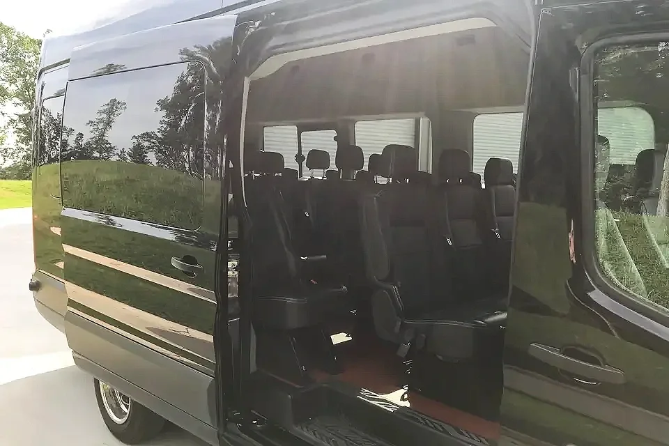 Black passenger van with sliding door open, showing multiple rows of seats inside, parked outdoors on a grassy area with trees in the background.