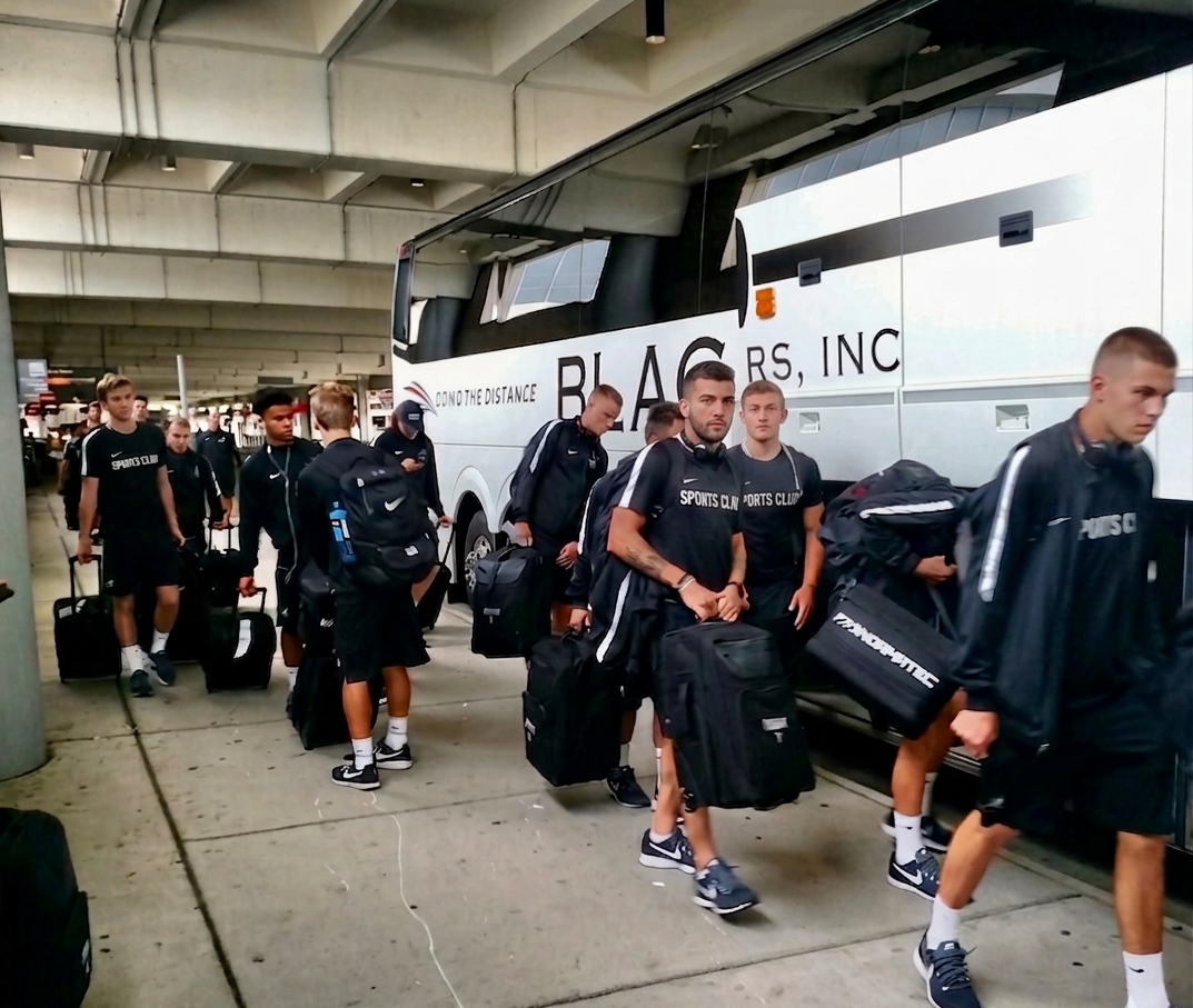 Group of young men in athletic clothing with backpacks and rolling suitcases boarding a bus at an airport.