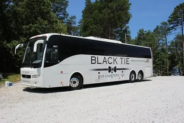White bus with black and white design, text reading "BLACK TIE" and "BUS CHARTERS, INC." parked on gravel with trees in the background.