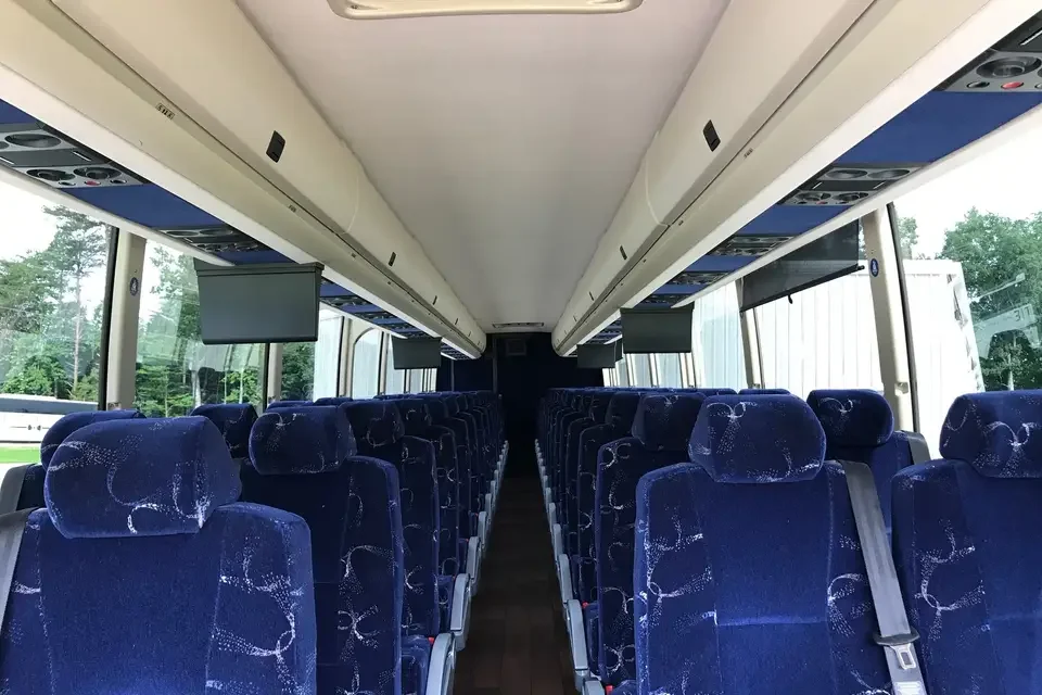 Interior of a bus with rows of blue fabric seats, overhead compartments, and multiple monitors hanging from the ceiling.