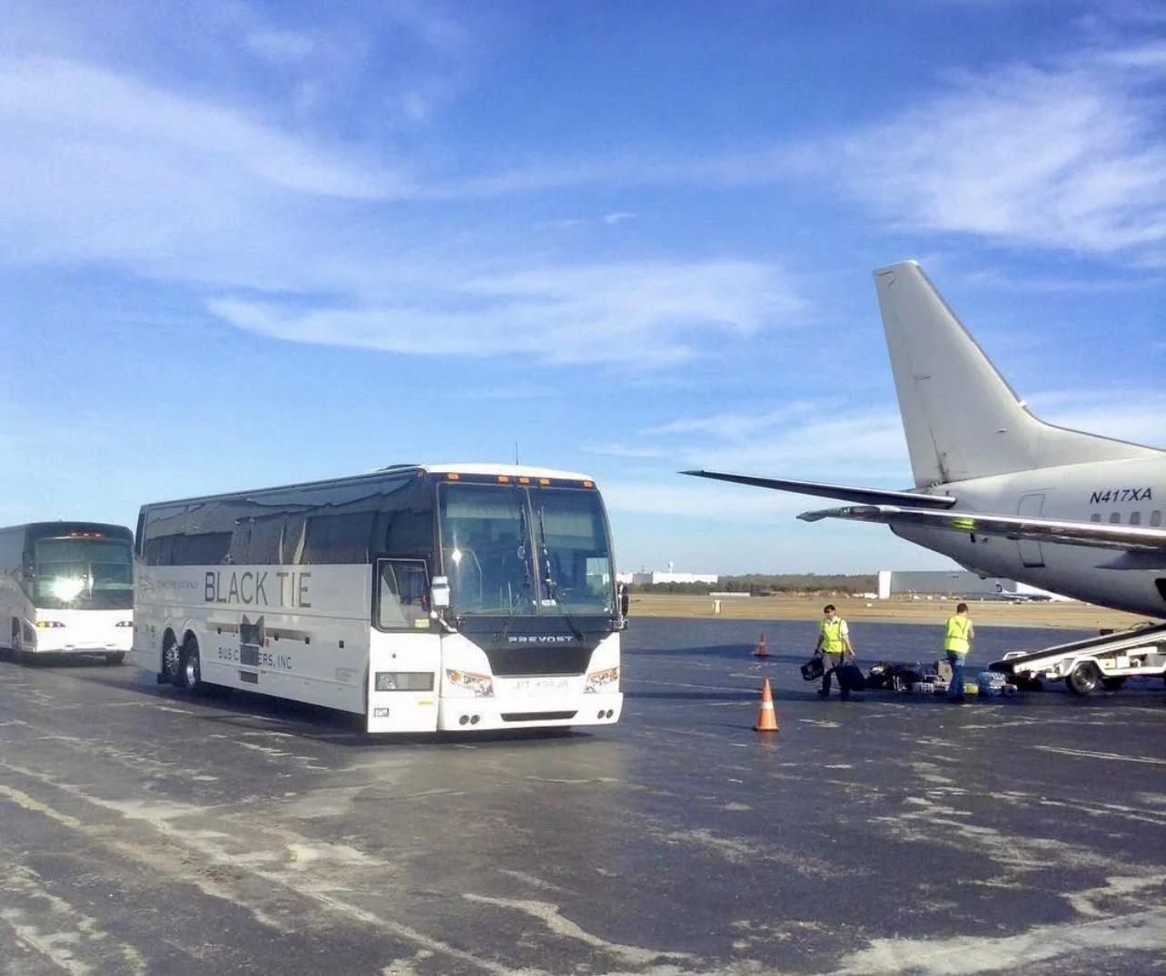 An airport tarmac with a bus labeled 'Black Tie' parked near an airplane, with two ground crew members in yellow vests handling luggage. Clear blue sky with some clouds in the background.