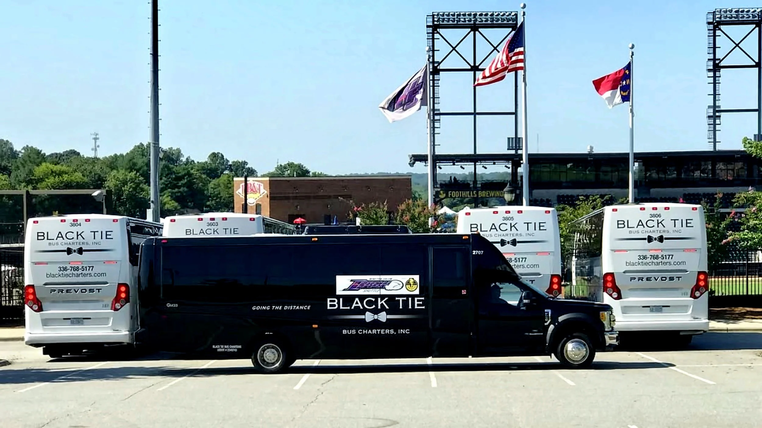 A parking lot with several buses branded with 'Black Tie Bus Charters, Inc.' and phone number, parked in front of a building with flags flying on poles, including the United States flag and flags with logos, and trees in the background.
