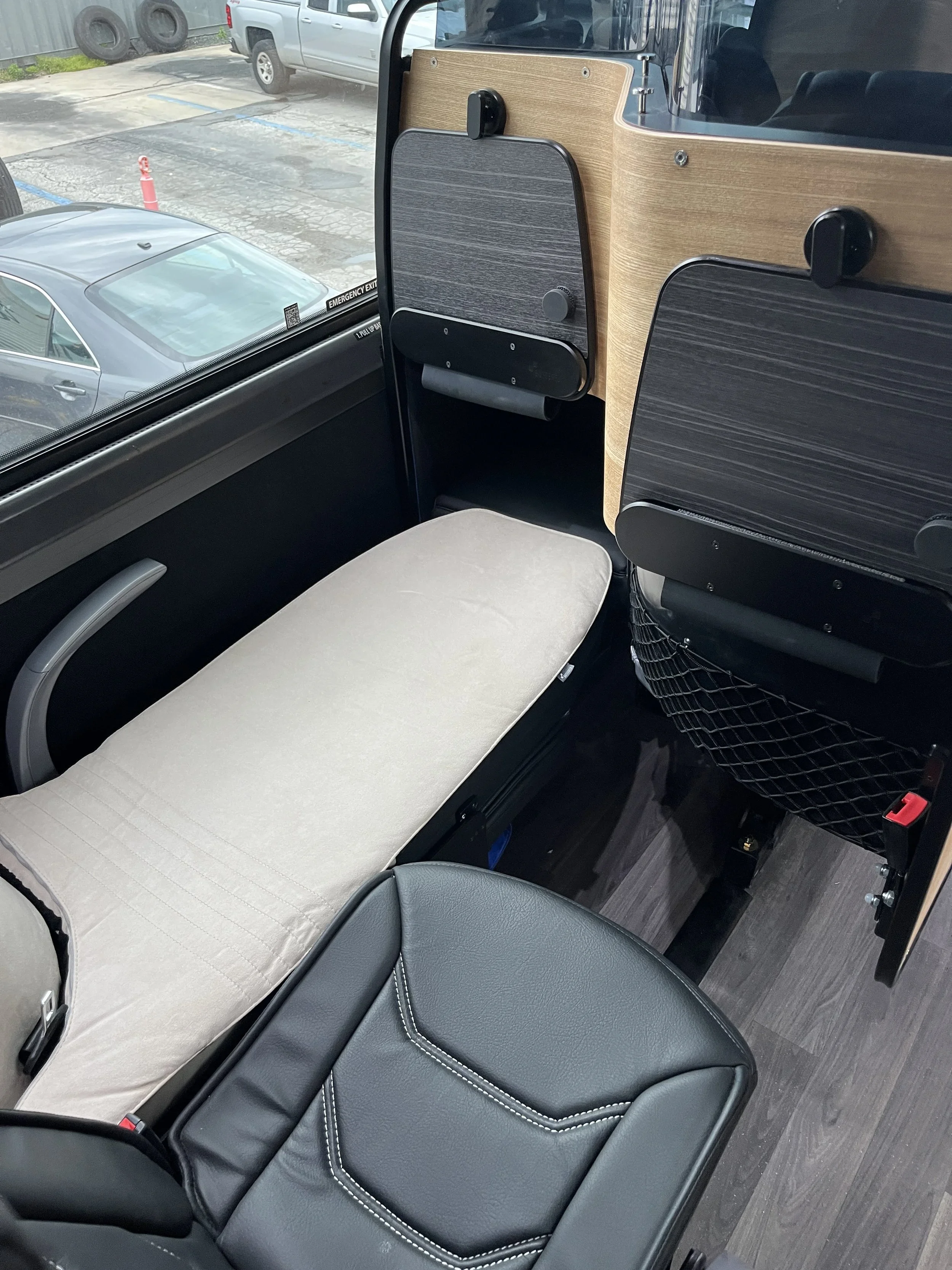 Interior view of a bus or train with a beige cushioned seat, black leather armrest, and wooden partition. Outside the window, parked cars and a parking lot are visible.