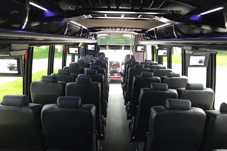 Interior of a modern bus with black leather seats, screens above the windows, and a view of green scenery outside.