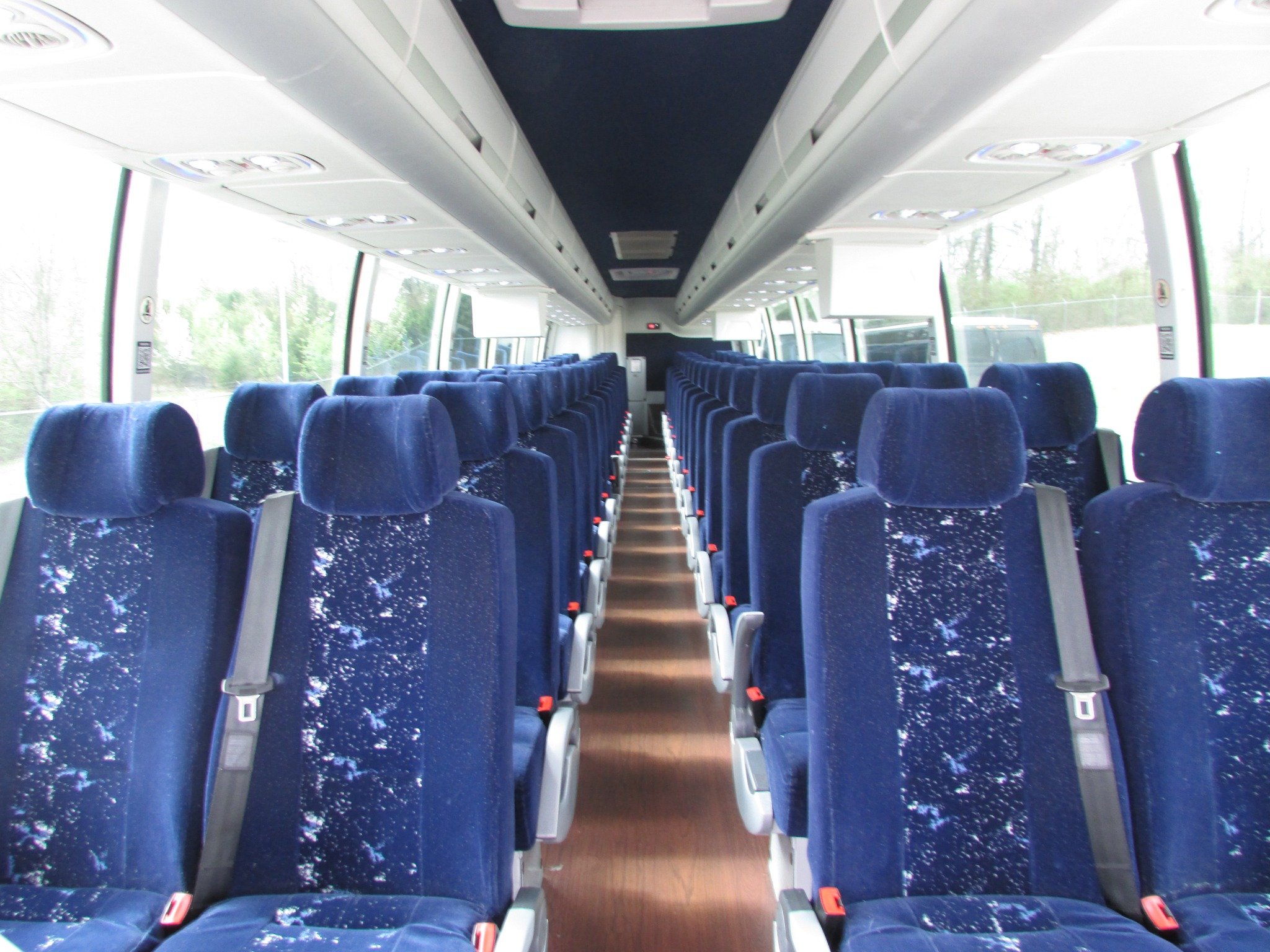 Empty bus interior with blue upholstered seats, overhead compartments, and large windows showing trees and sky outside.