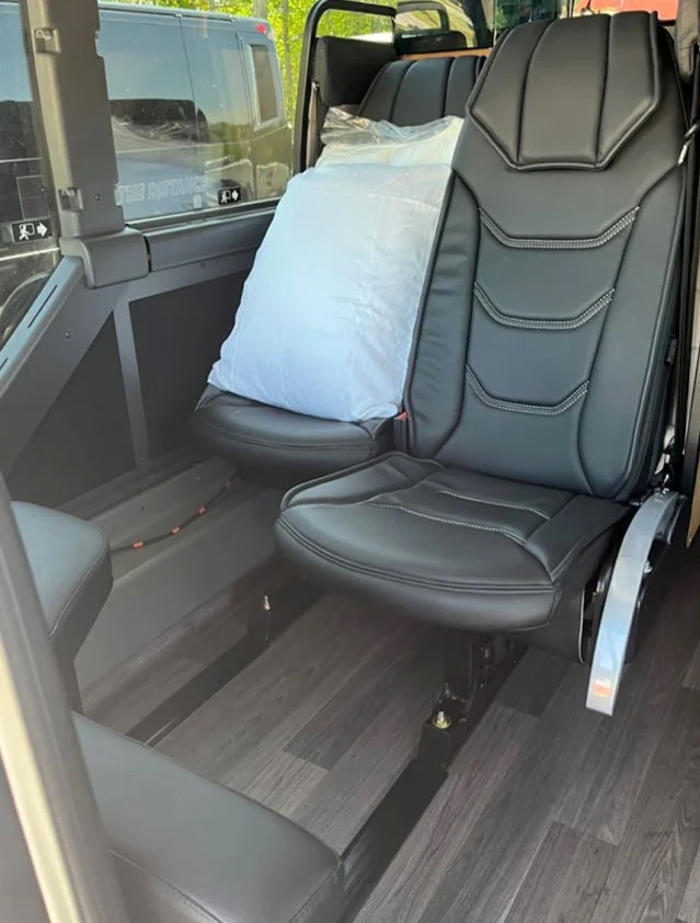 Inside a helicopter, showing one closed leather seat and one seat with a pillow, with large windows and a view of a vehicle outside.