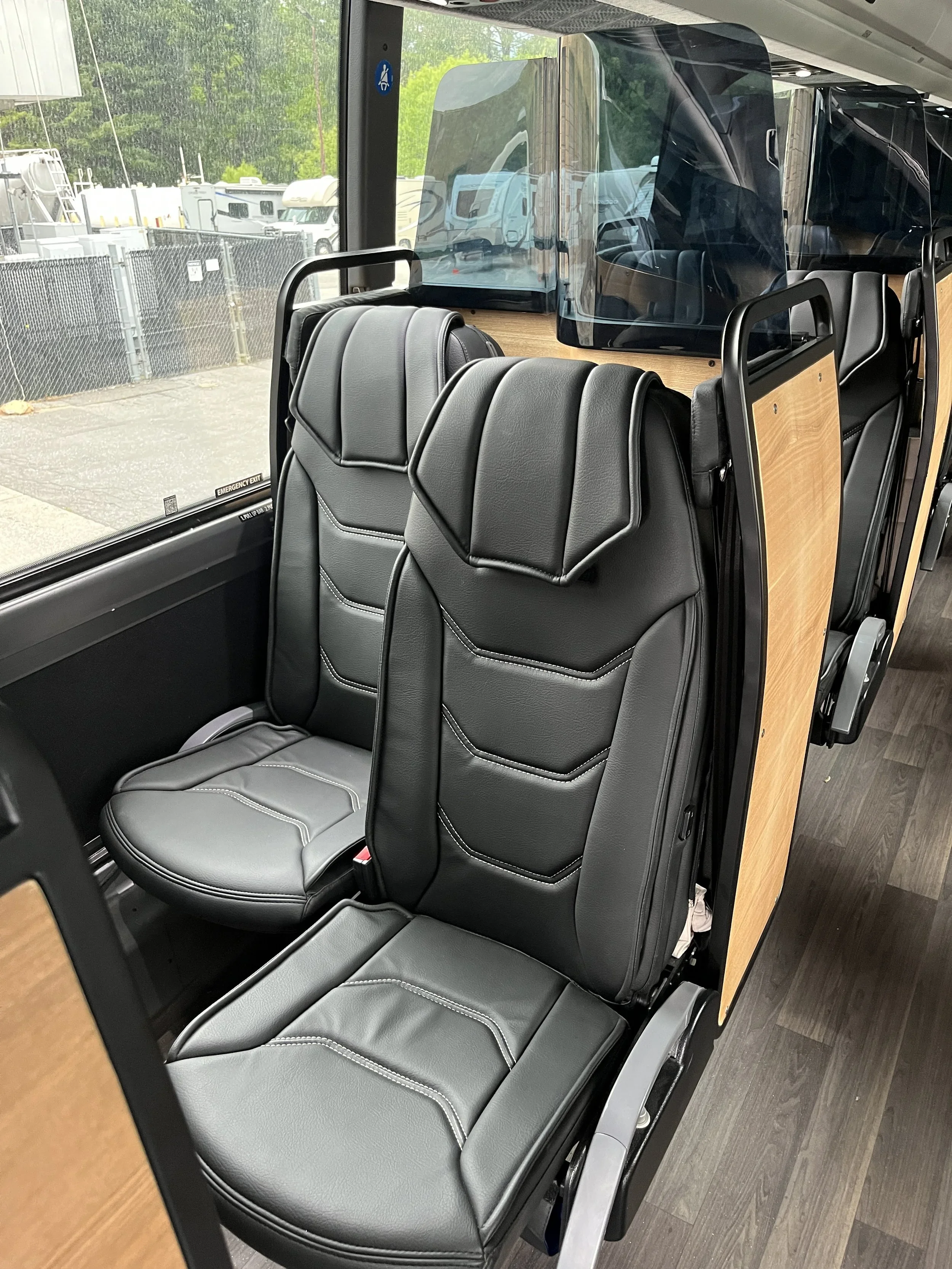 Interior of a bus with black leather seats and wood paneling on the sides, looking out the window at parked RVs and a chain-link fence outside.