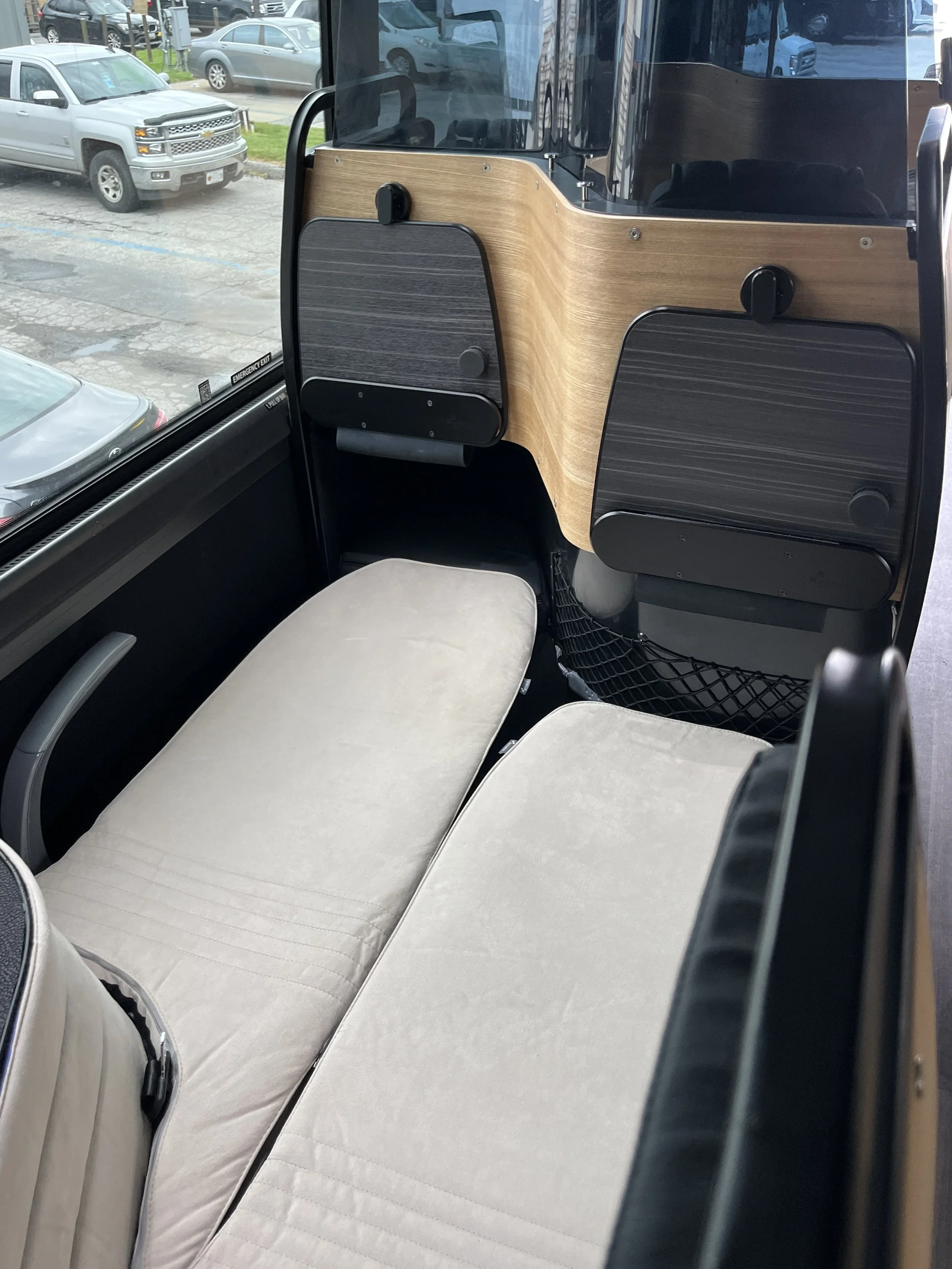 Interior of a bus with a beige cushioned seat and a wooden partition. Outside the window, parking lot with silver trucks and cars are visible.