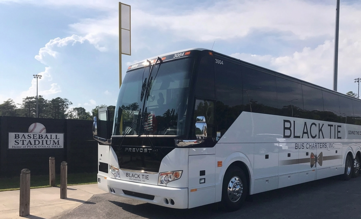 White and black bus with 'Black Tie Bus Charters, Inc.' written on the side, parked near a baseball stadium with a sign that says 'Baseball Stadium Home of Puck O'Neal Field,' under a partly cloudy sky.