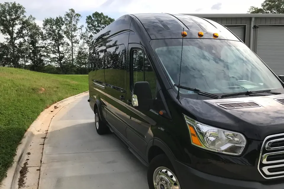 A black passenger van parked on a paved driveway next to a grassy area with trees in the background.