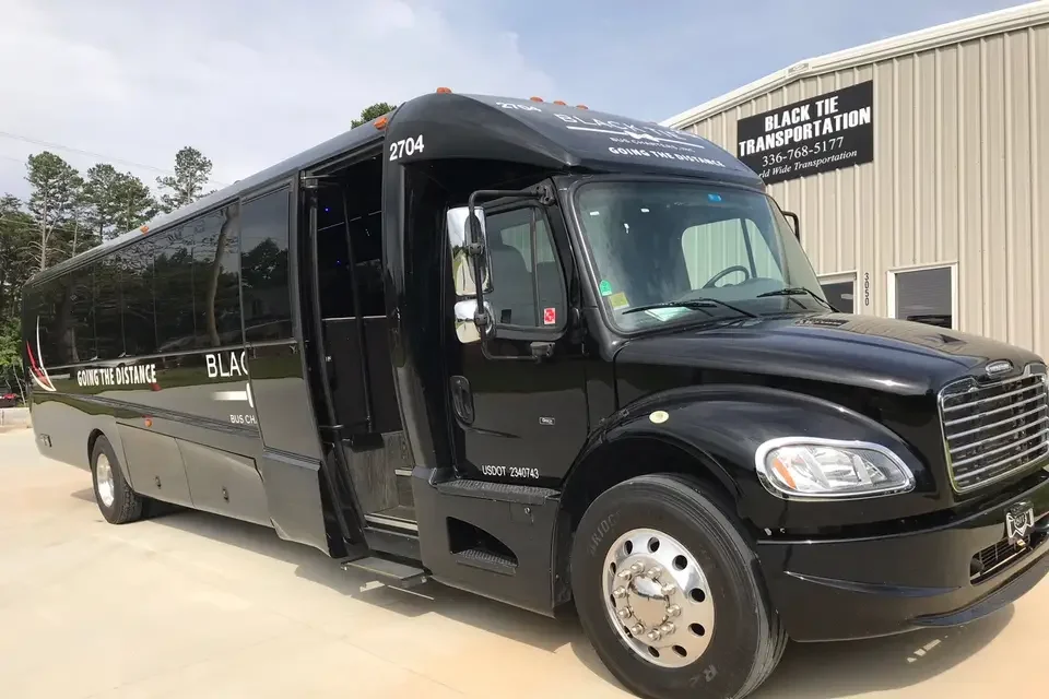 Black bus parked outside a beige commercial building with a sign that reads "Black Tie Transportation" and a phone number.