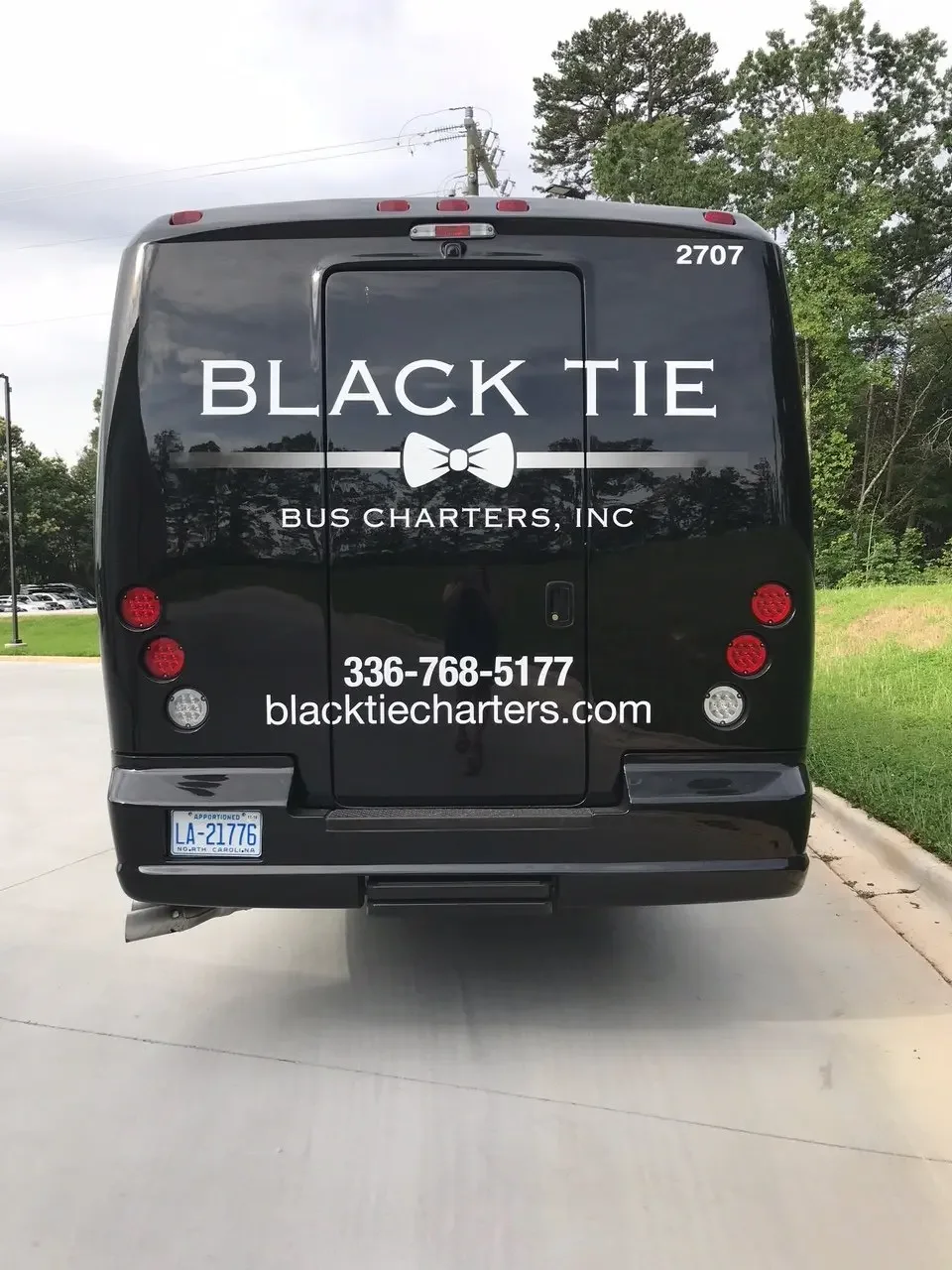 Black bus with company logo and contact information, parked on a concrete parking lot with trees and cloudy sky in the background.