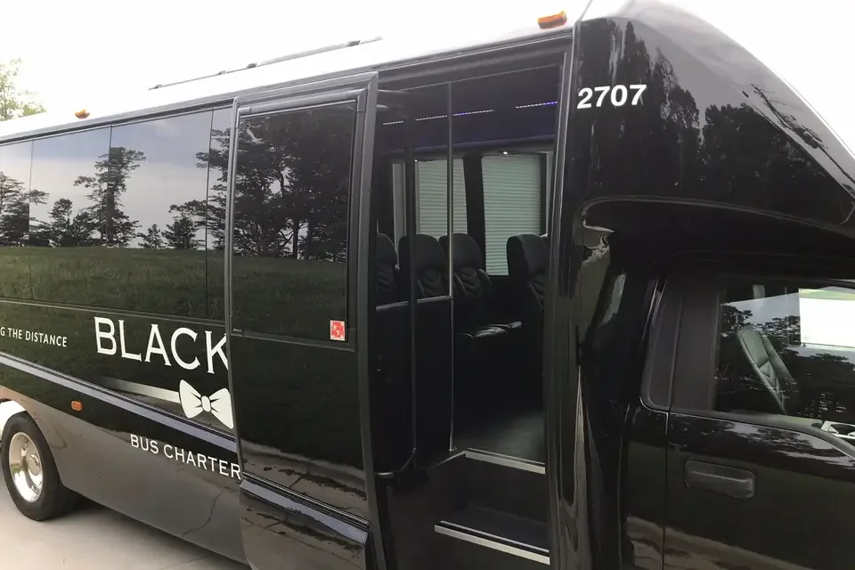 Black bus with large tinted windows and open door, parked outdoors with trees in the background. The bus has the words 'Black' and 'Bus Charter' visible, along with a bowtie logo and a slogan suggesting maintaining distance.