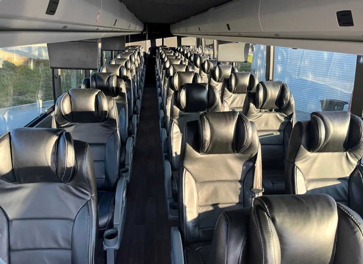 Interior of a bus with leather seats arranged in rows facing forward, with seatbelts visible and large windows on each side.