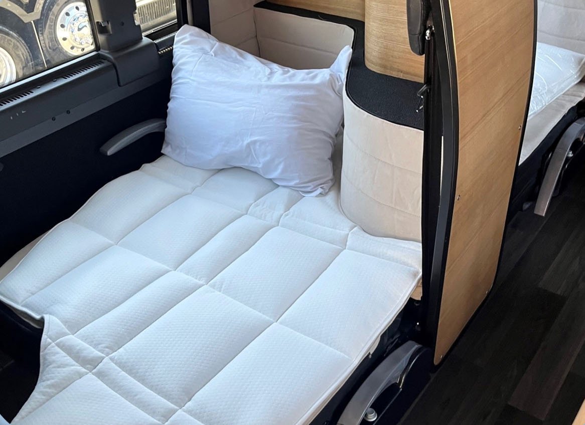 Interior view of a sleeper cab with a bed laid out with a white quilt and pillow, adjacent to a wooden partition, inside a vehicle.