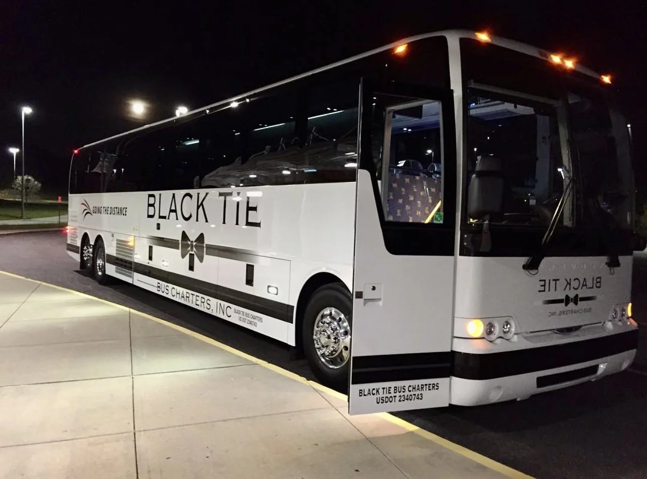 A white bus parked at night with 'Black Tie Bus Charters' written on the side and top, featuring a small bow tie graphic and door open showing patterned seats inside.