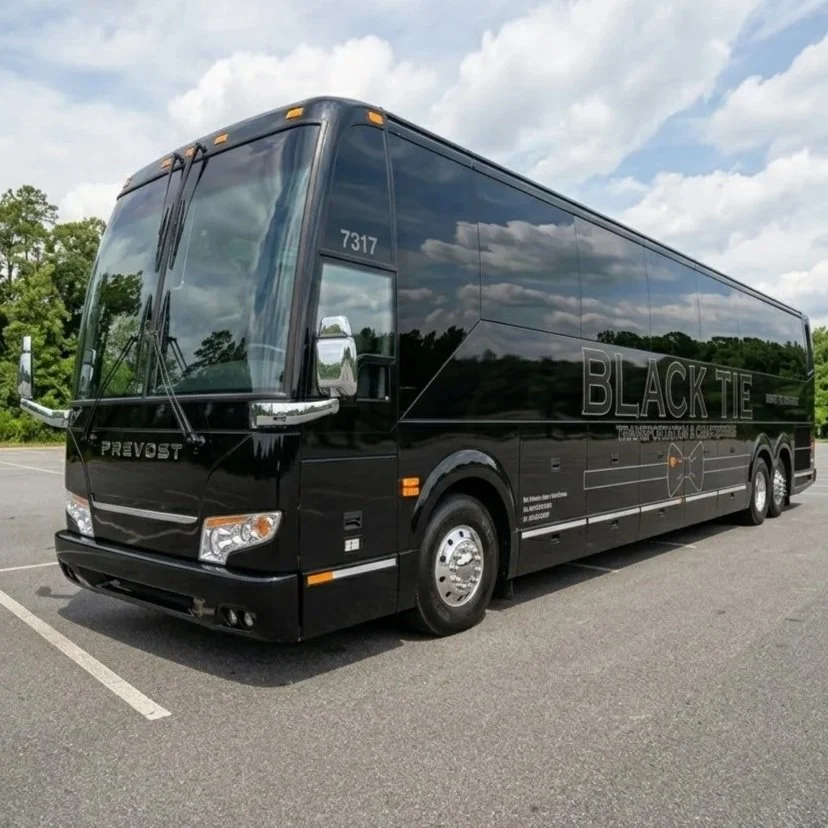 Black tour bus with the words 'BLACK TIE' on the side, parked in an empty lot with green trees and a partly cloudy sky in the background.