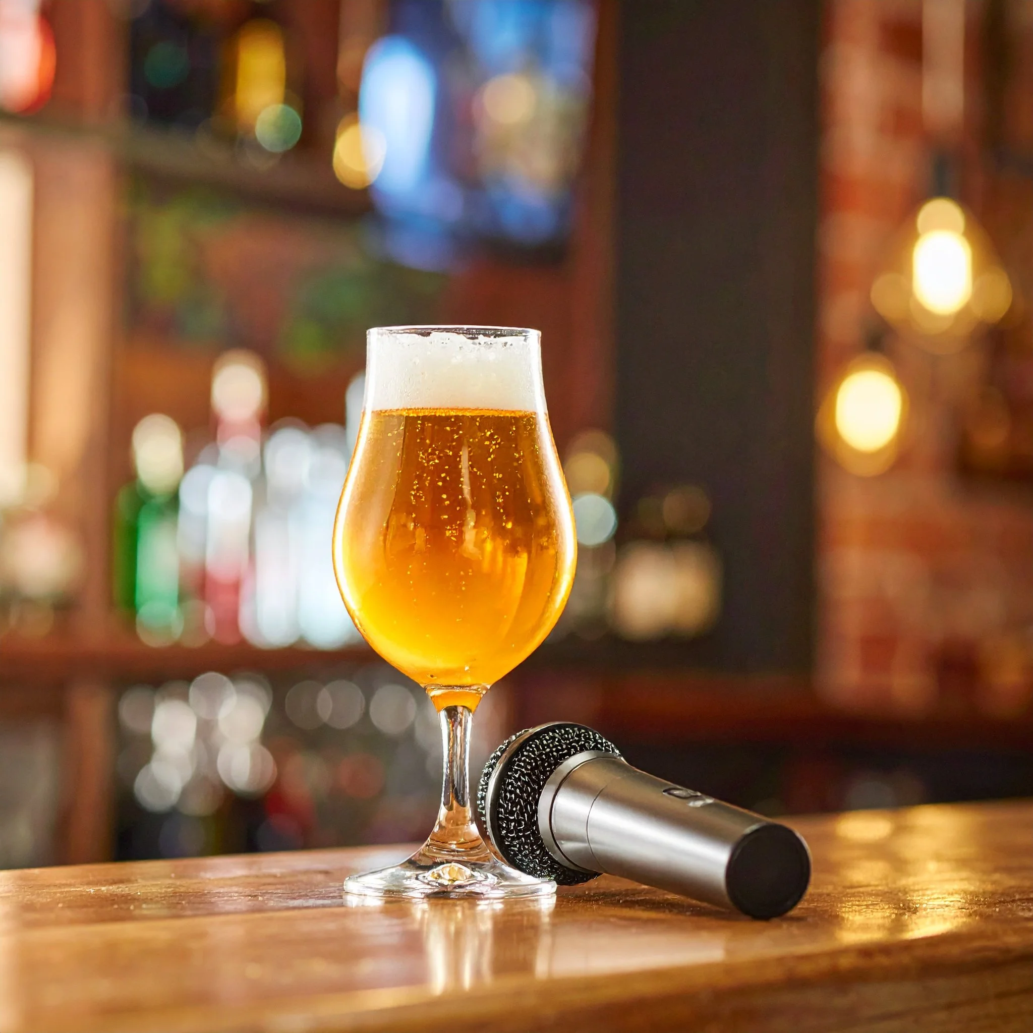 A glass of beer on a wooden bar counter with a beer cap and a metallic object nearby, blurred colorful lights in the background.