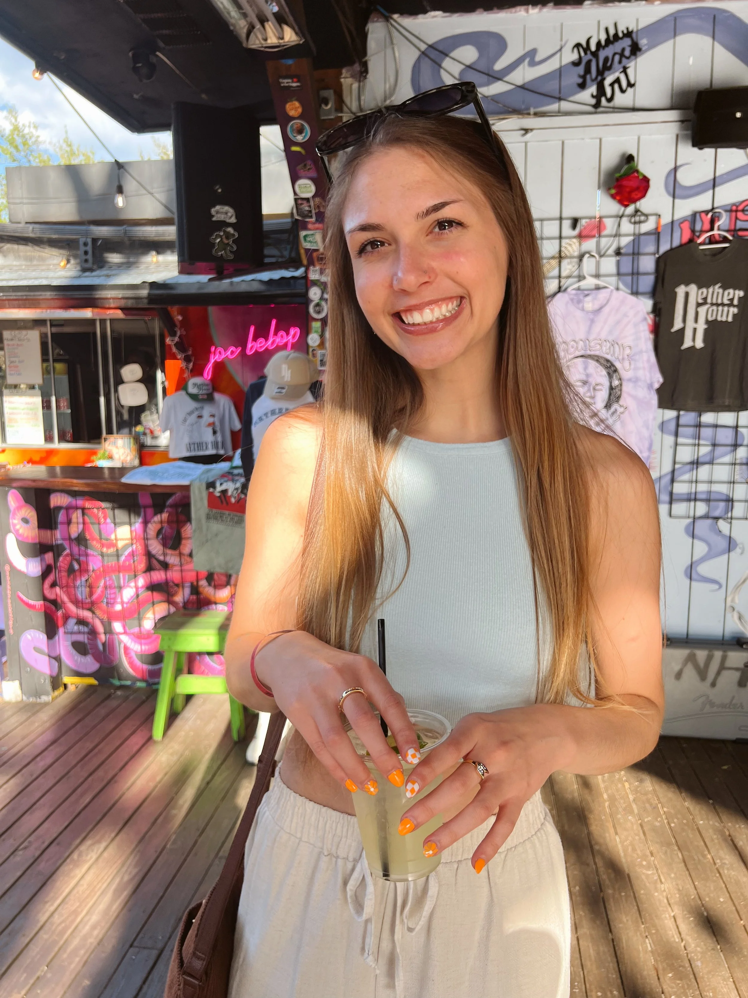 A woman with long light brown hair and sunglasses on her head, smiling and holding a drink with a straw at an outdoor location with a wooden floor and merchandise shop in the background.