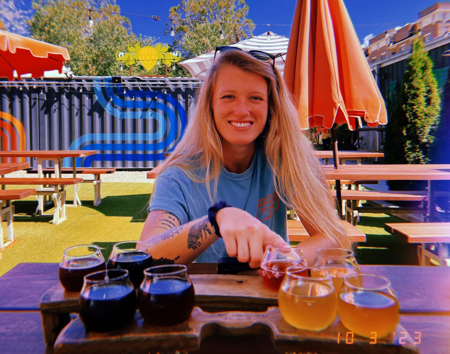 A smiling woman with long blonde hair sitting at an outdoor table with tasting glasses of beer in front of her. She is pointing at one of the glasses. There are orange umbrellas and wooden picnic tables in the background, along with a colorful mural and string lights in a sunny setting.