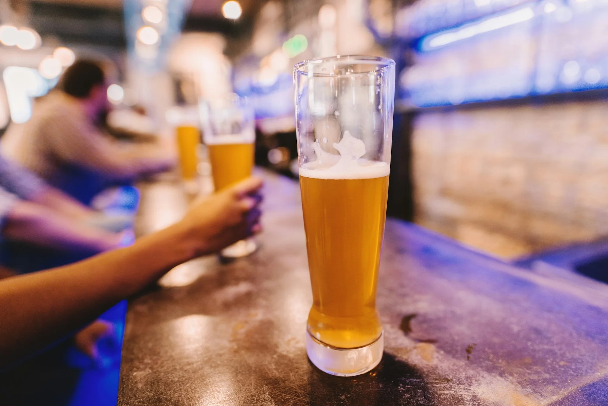 Close-up of a tall, slender glass filled with amber-colored beer on a bar counter with blurred patrons in the background.