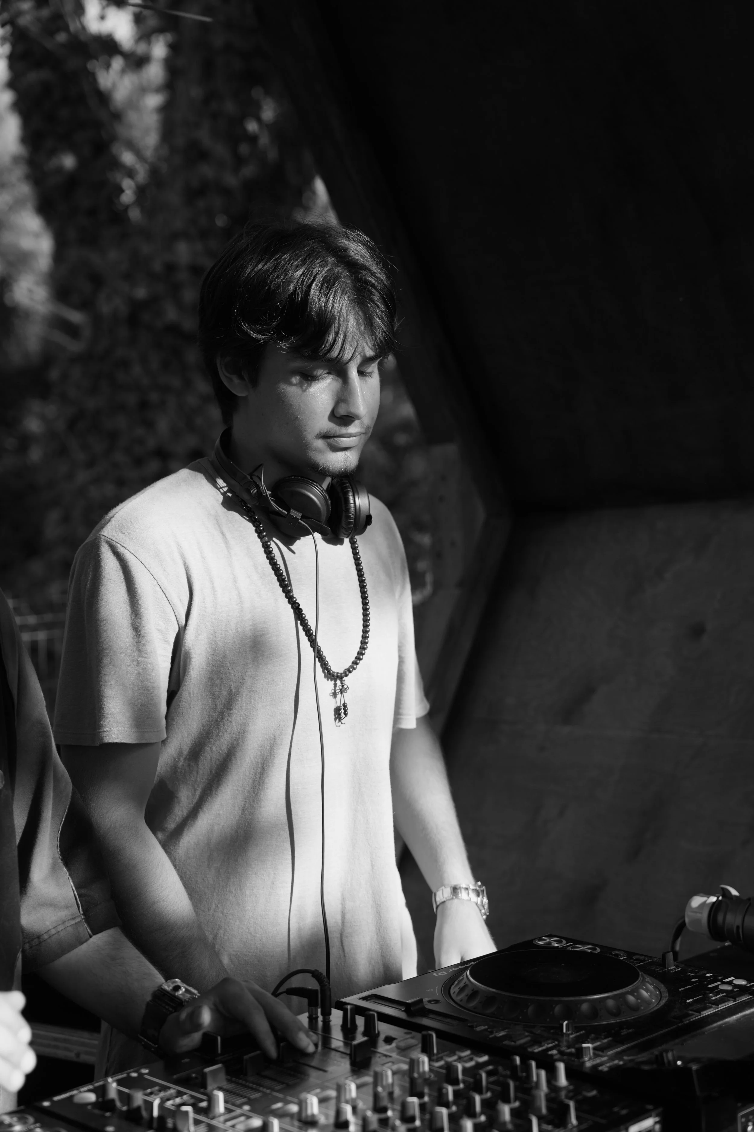 A young man with headphones around his neck is DJing with a turntable and mixer, wearing a t-shirt, wristwatch, and beaded necklace, in an outdoor setting.