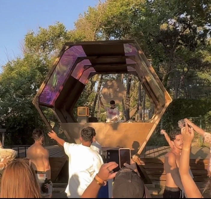 People dancing and taking photos in front of a large hexagonal wooden and glass DJ booth outdoors during sunset.