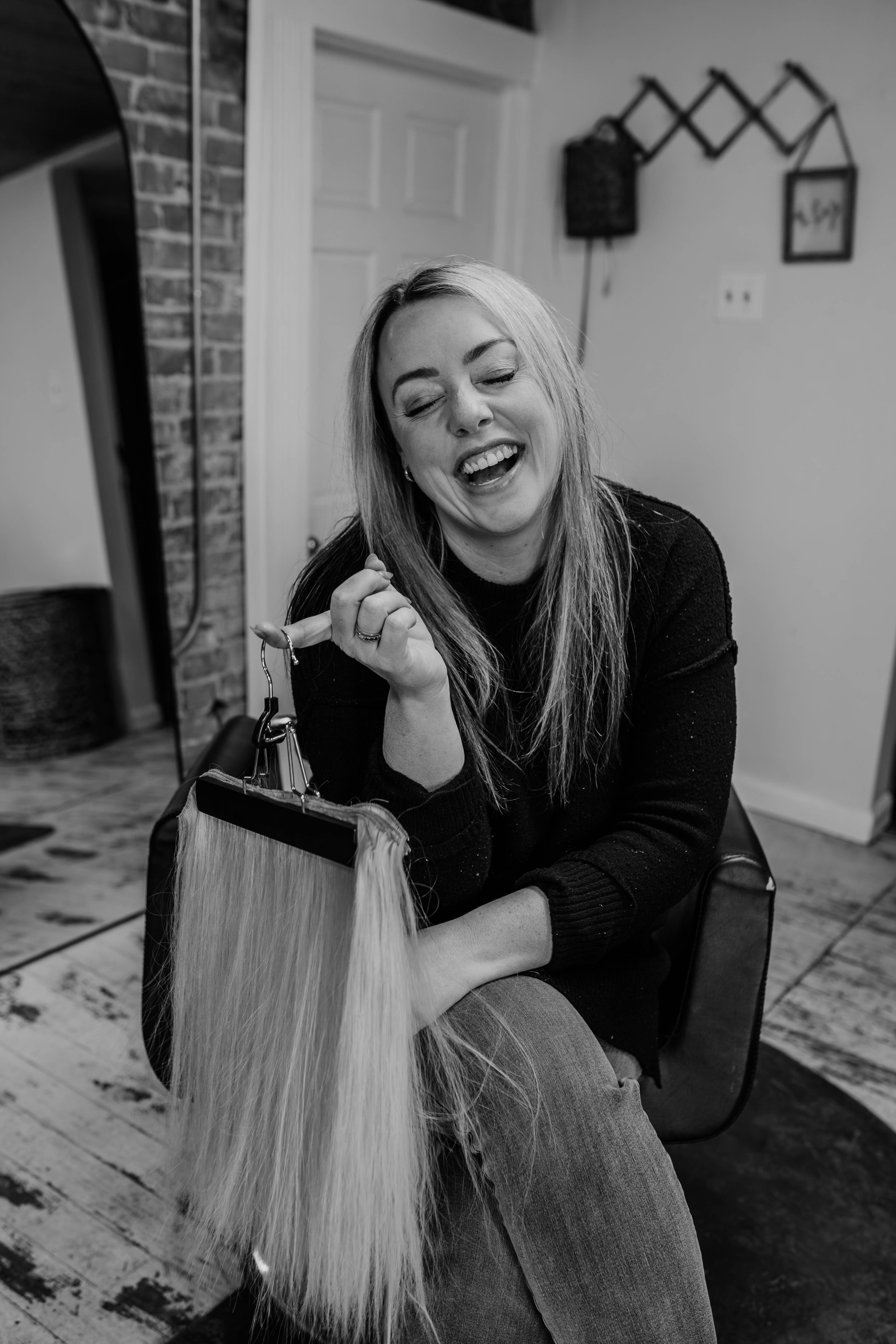 A smiling woman with long blonde hair sitting indoors, holding a set of keys and a long wig of blonde hair. She is wearing a black top and light jeans. The background features a brick wall, a white door, a coat rack, and framed pictures.