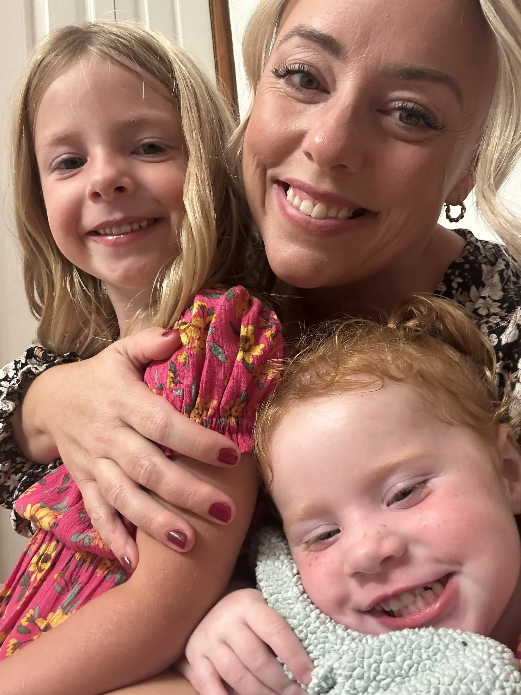 A smiling woman with blonde hair and earrings hugging a young girl with blonde hair and freckles, and another smiling child with red hair.