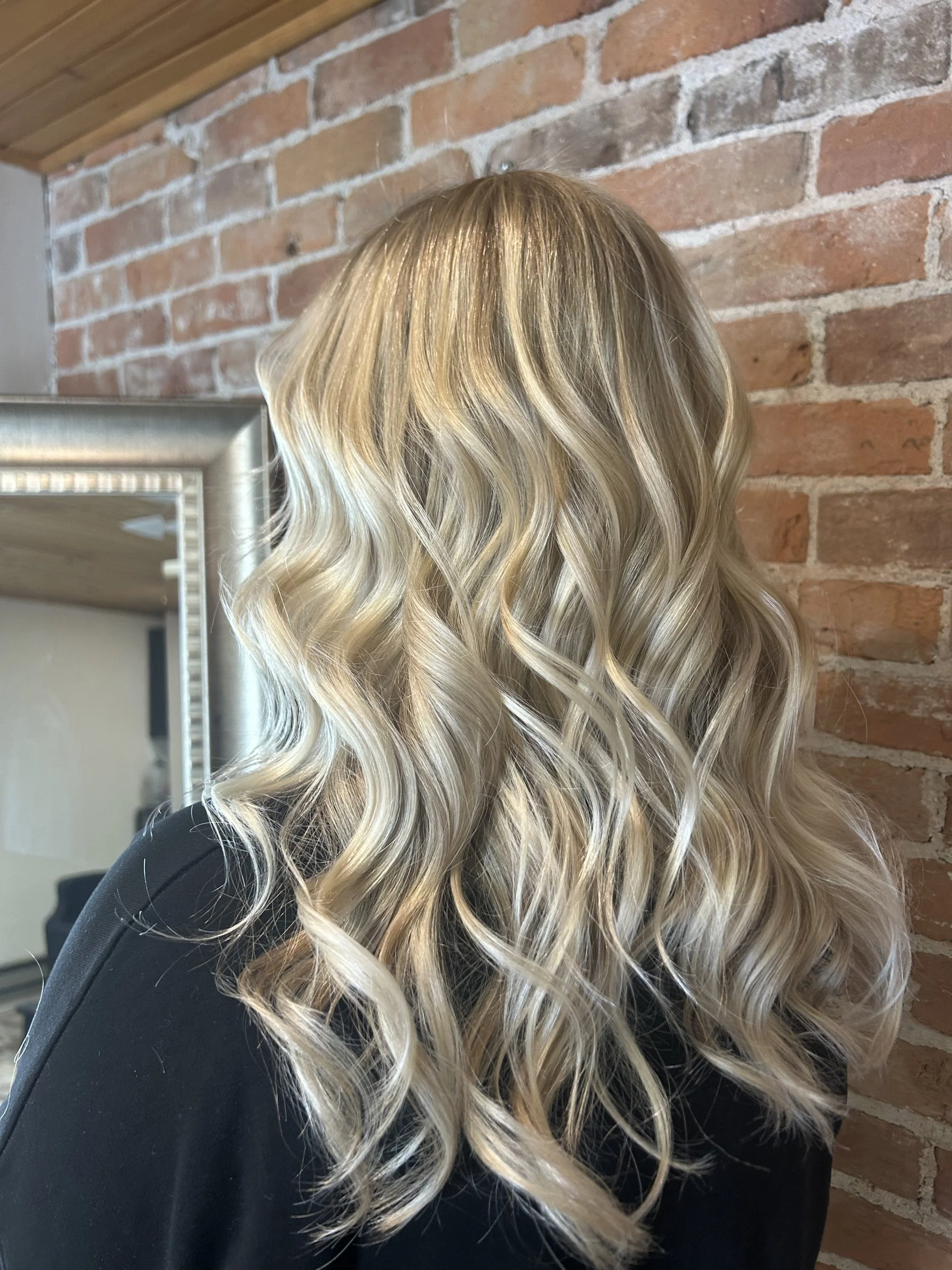 Person with long, curly, blonde hair standing in front of a brick wall, wearing a black top.