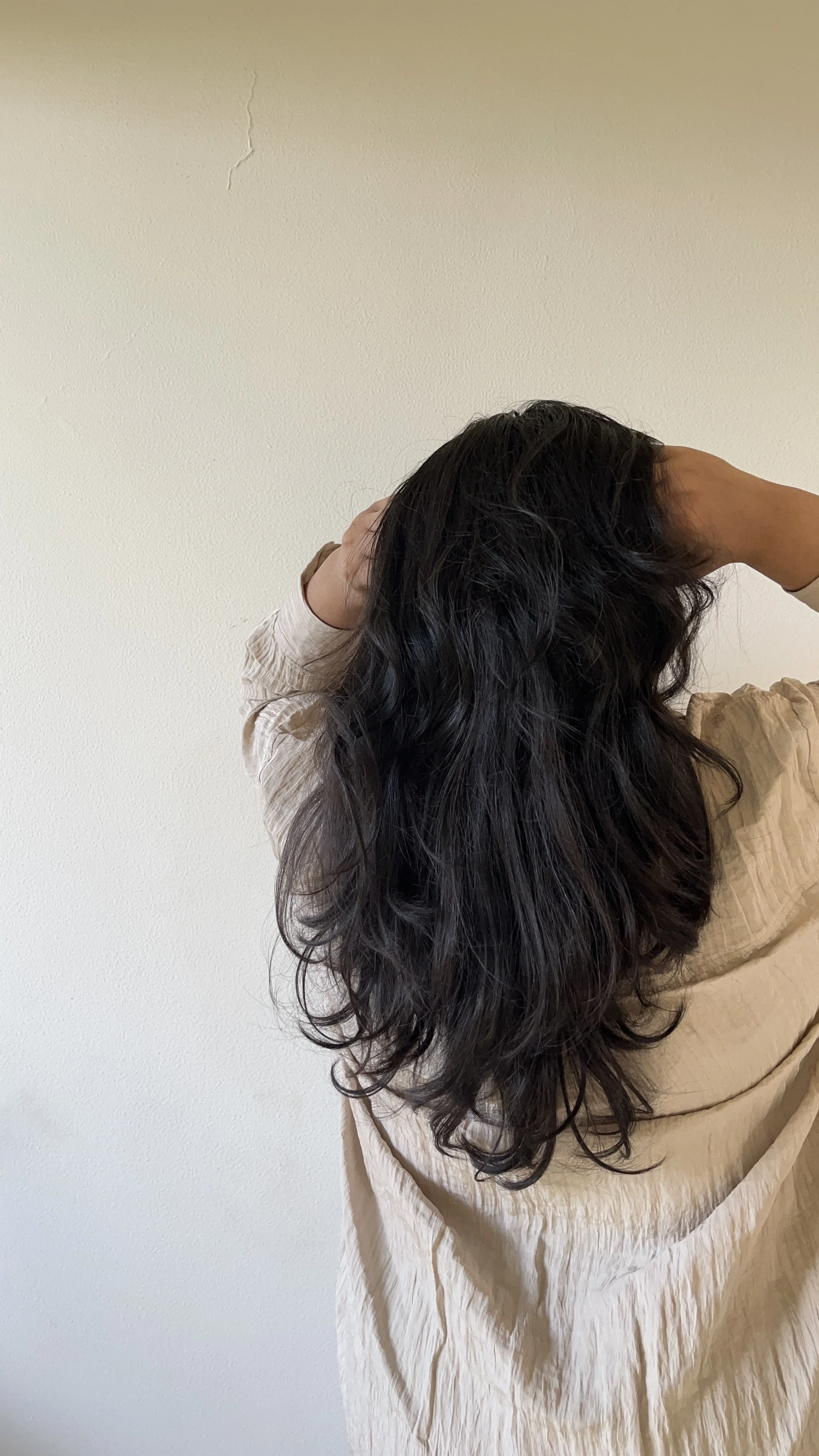 A person with long, wavy dark hair wearing a light-colored, loose dress, standing against a plain light-colored wall and holding their head with both hands.