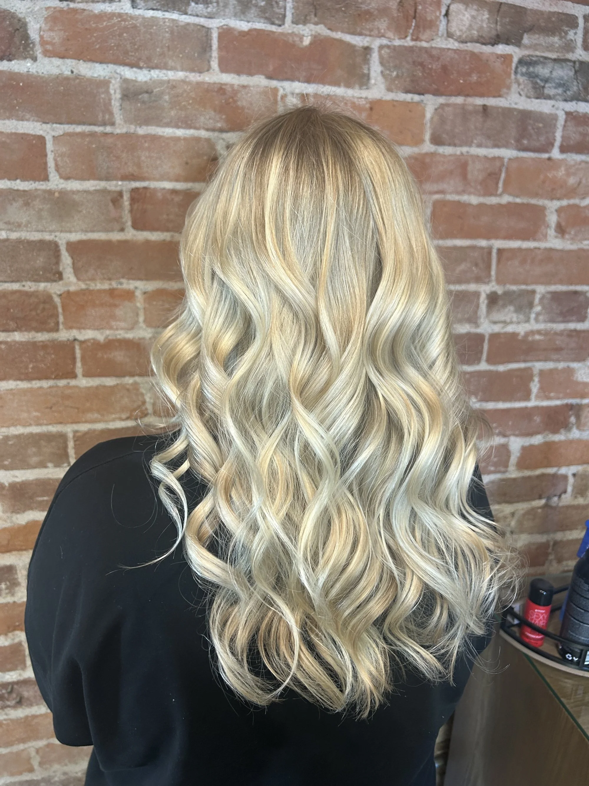 Back of a person with long, wavy blonde hair against an exposed brick wall, wearing a black top.
