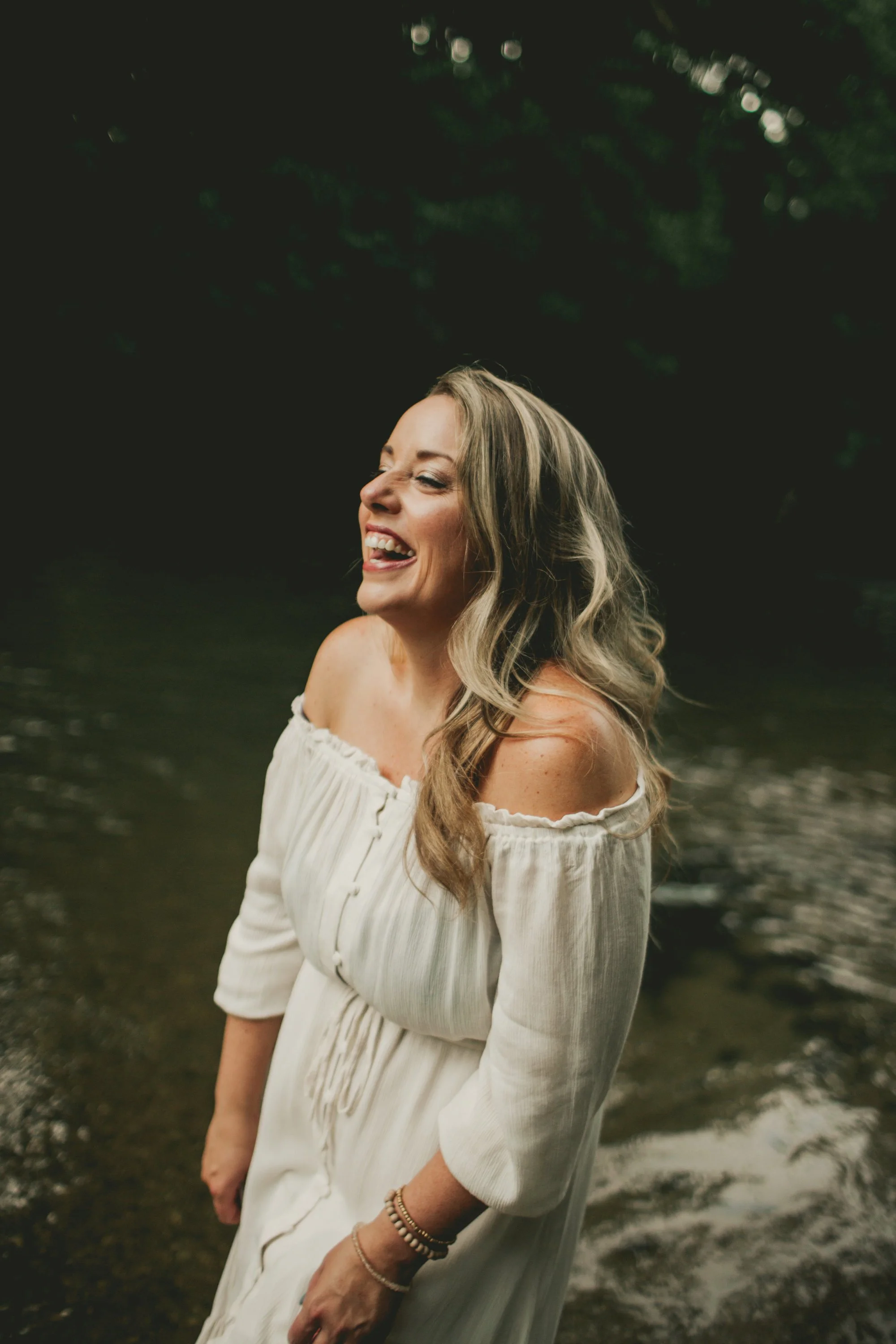 A woman with long, wavy blonde hair is smiling and sticking her tongue out while standing near a body of water in a natural outdoor setting during daytime.