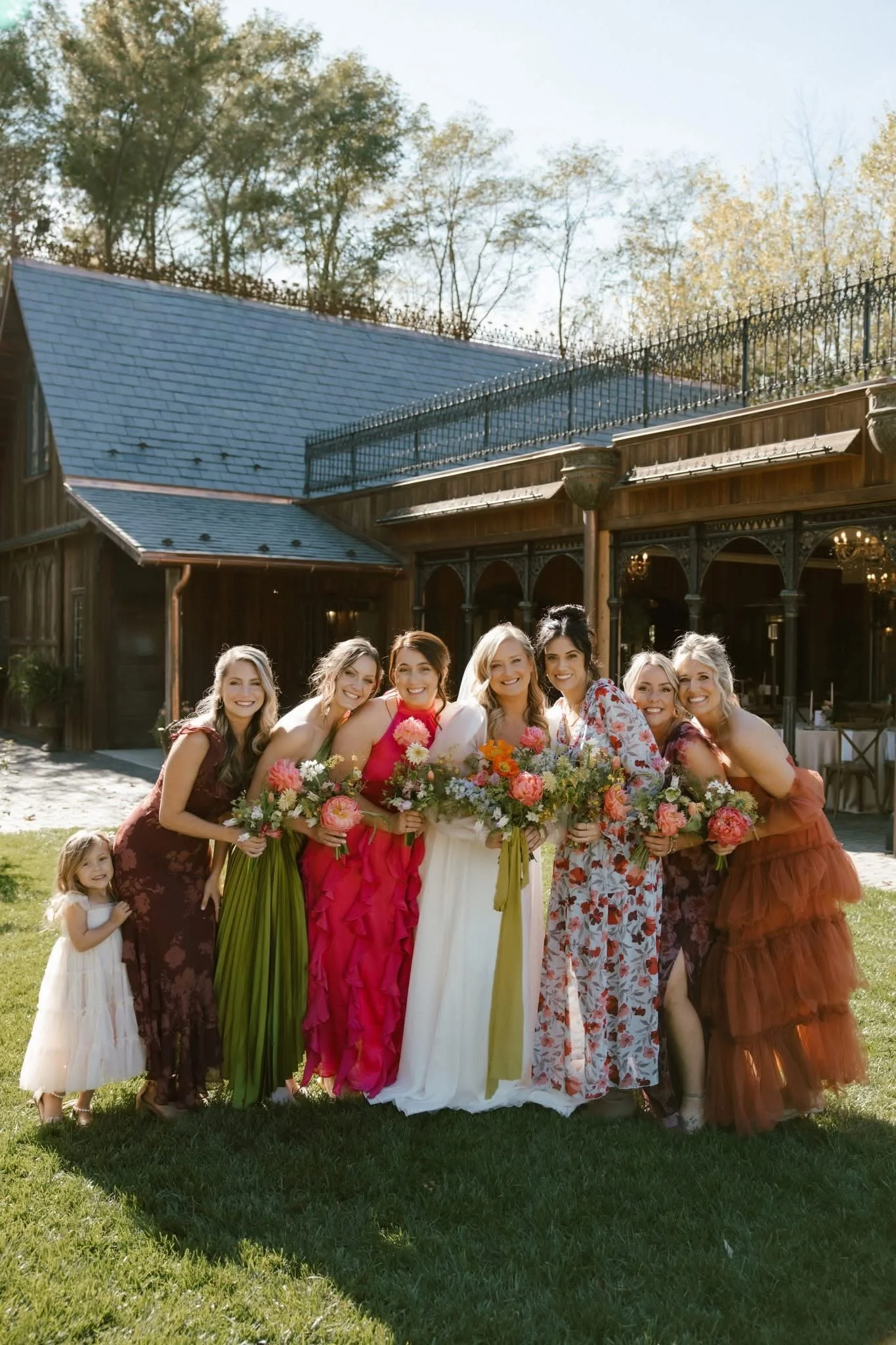 A diverse group of women and children in colorful dresses, holding bouquets of flowers, standing outdoors in front of a rustic building with a deck and a blue roof, during daytime.