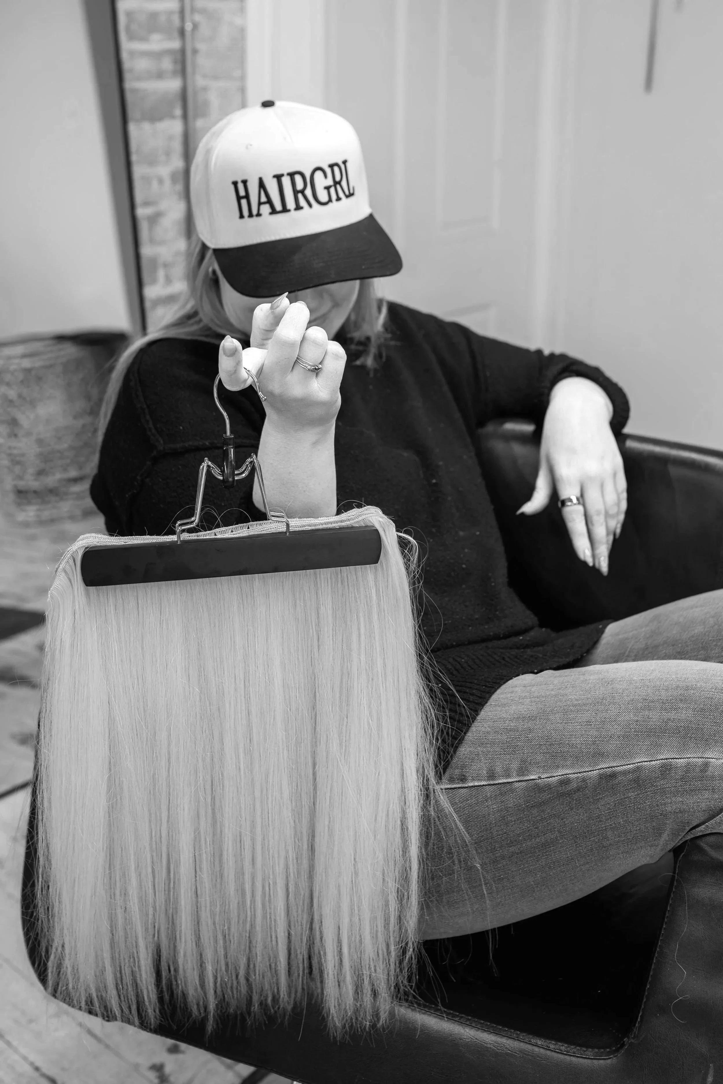 A person sitting on a chair, wearing a white baseball cap with the word 'HAIRGRL' written on it, and holding a wig on a stand in front of them.