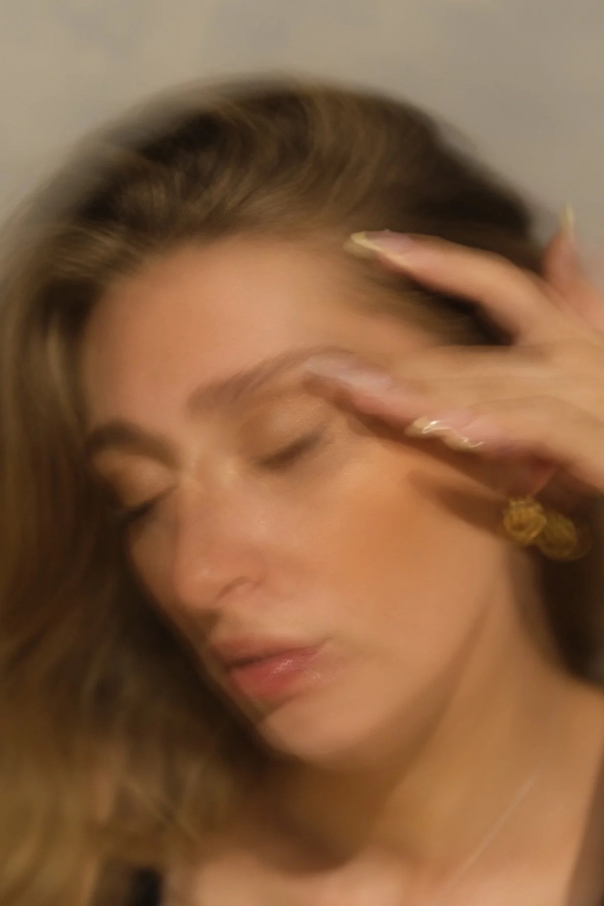 A woman with long brown hair and gold earrings, touching her face with her hand, eyes closed, with a neutral facial expression