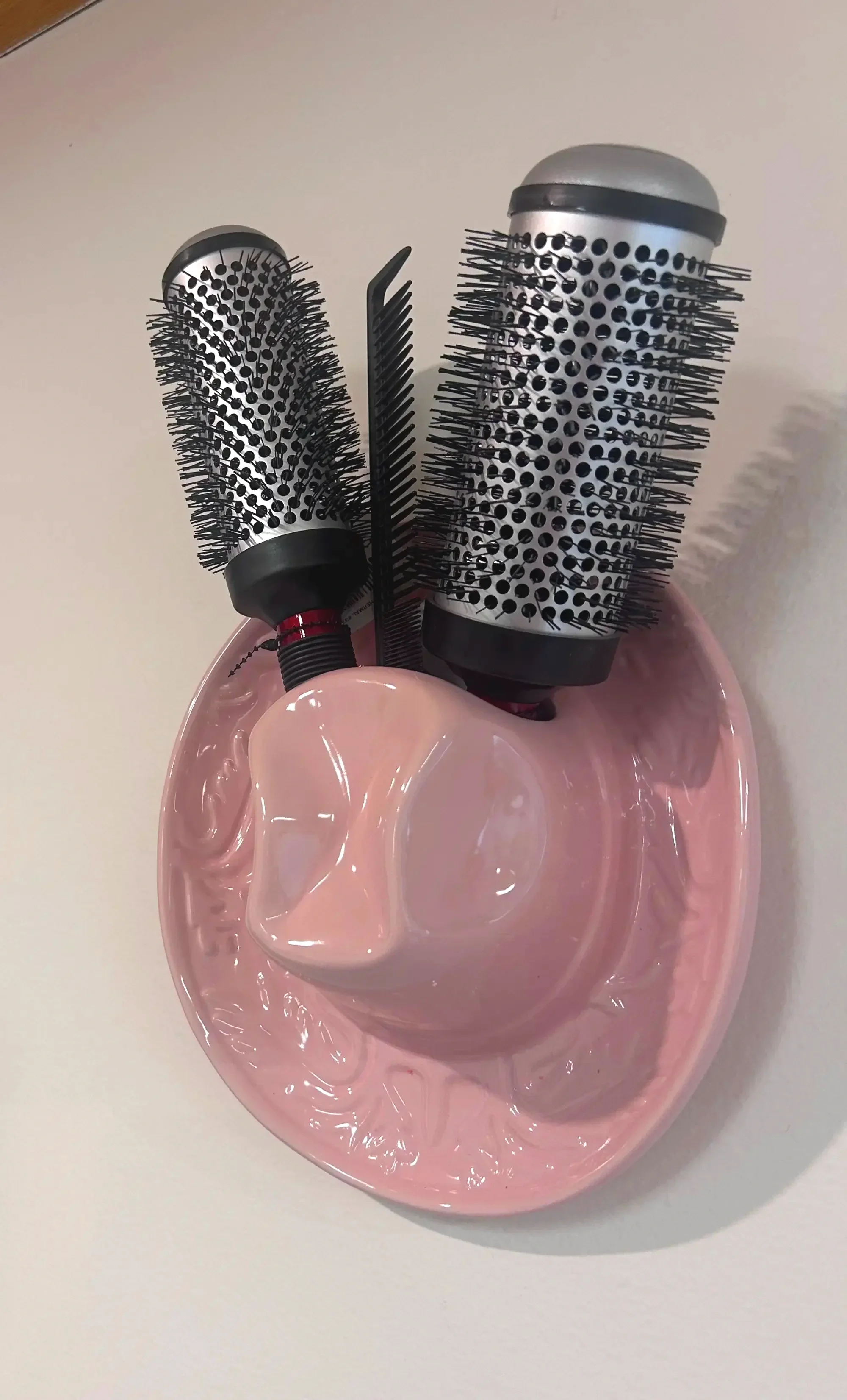 Two cylindrical hairbrushes with black bristles and a small black comb on a pink ceramic dish.