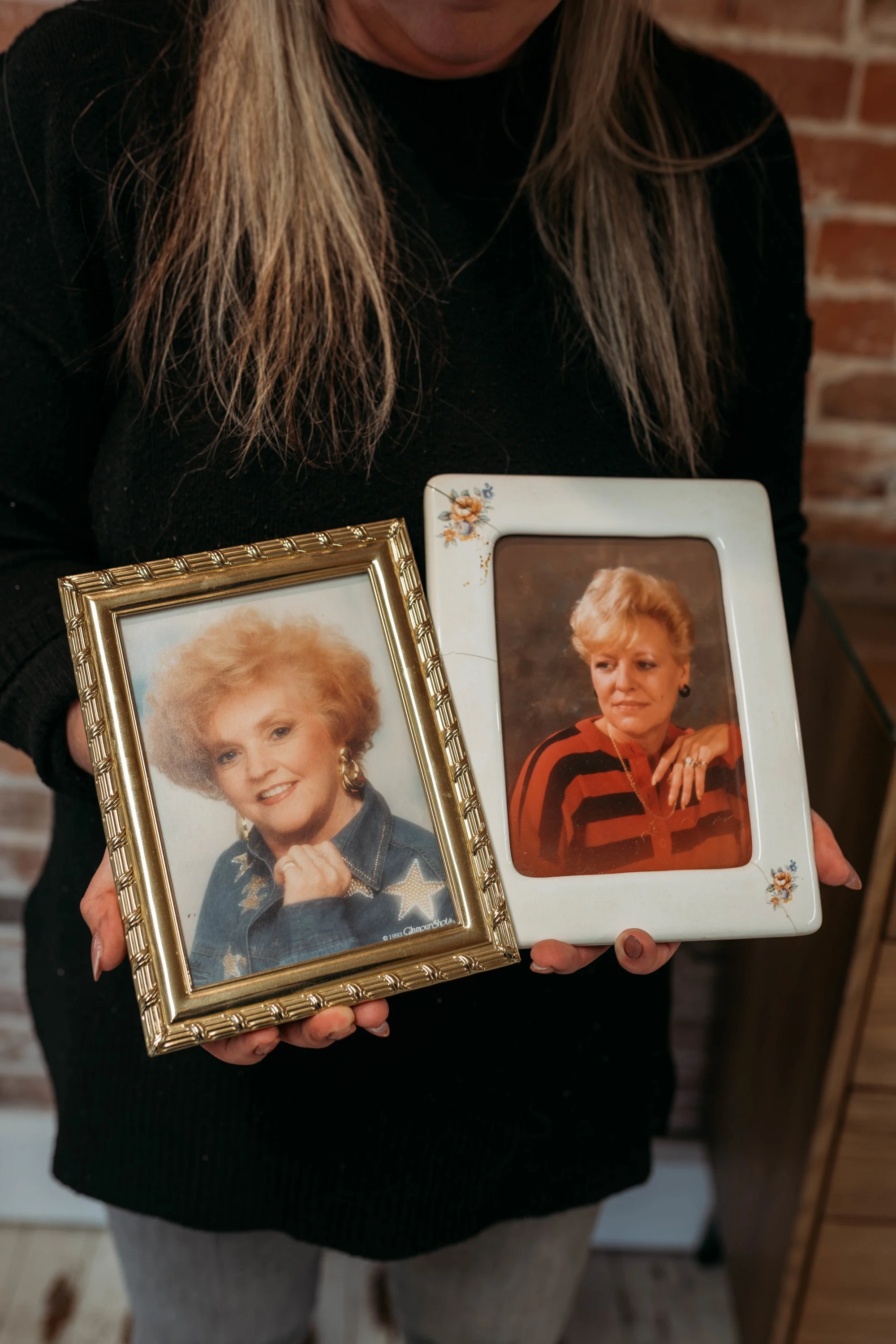 Person holding two framed photographs of women, one with a vintage hairstyle and denim jacket, the other with short blonde hair and a striped shirt. Brick wall in background.