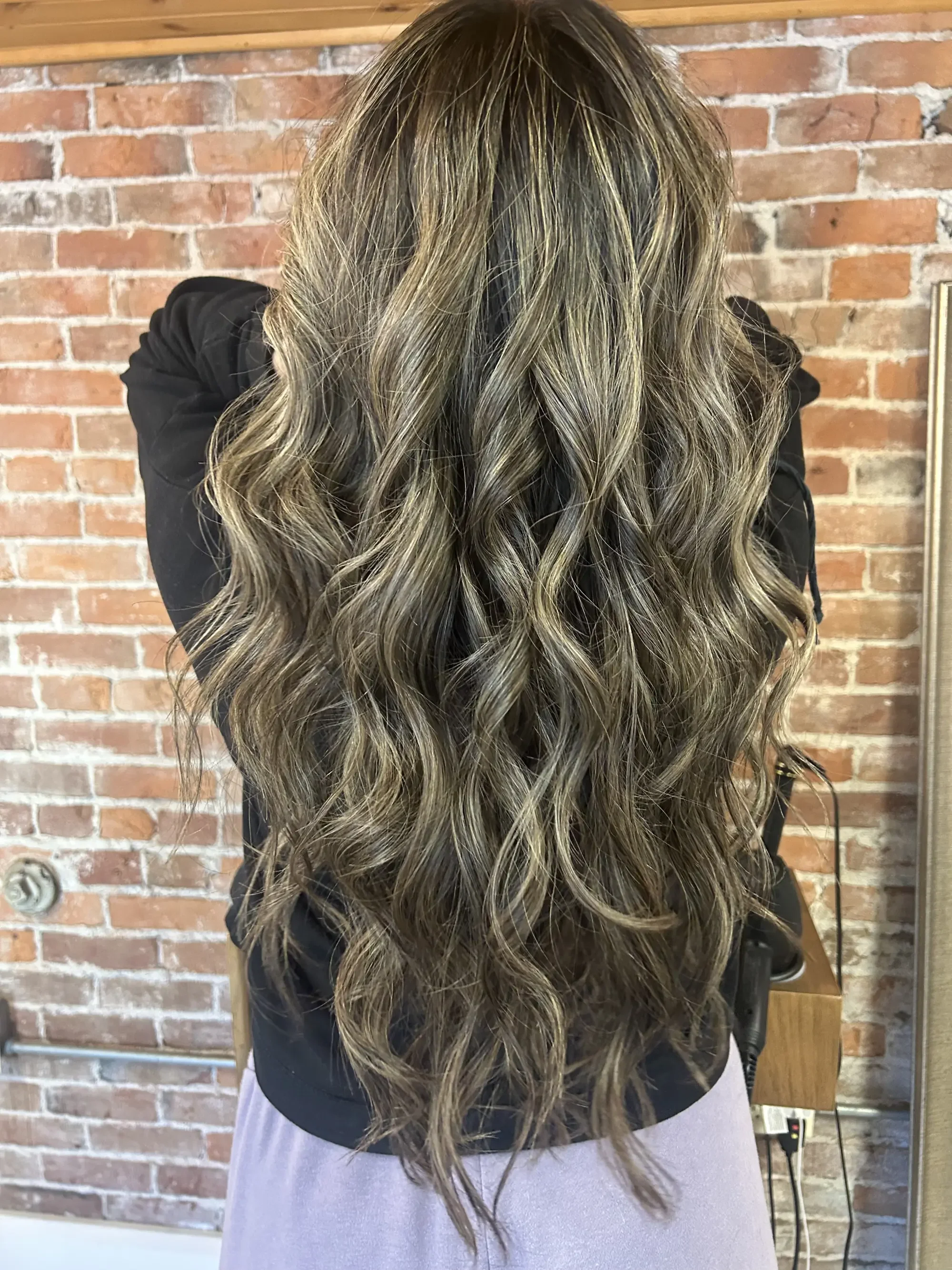 A woman with long, wavy hair with highlights, standing in front of a brick wall.