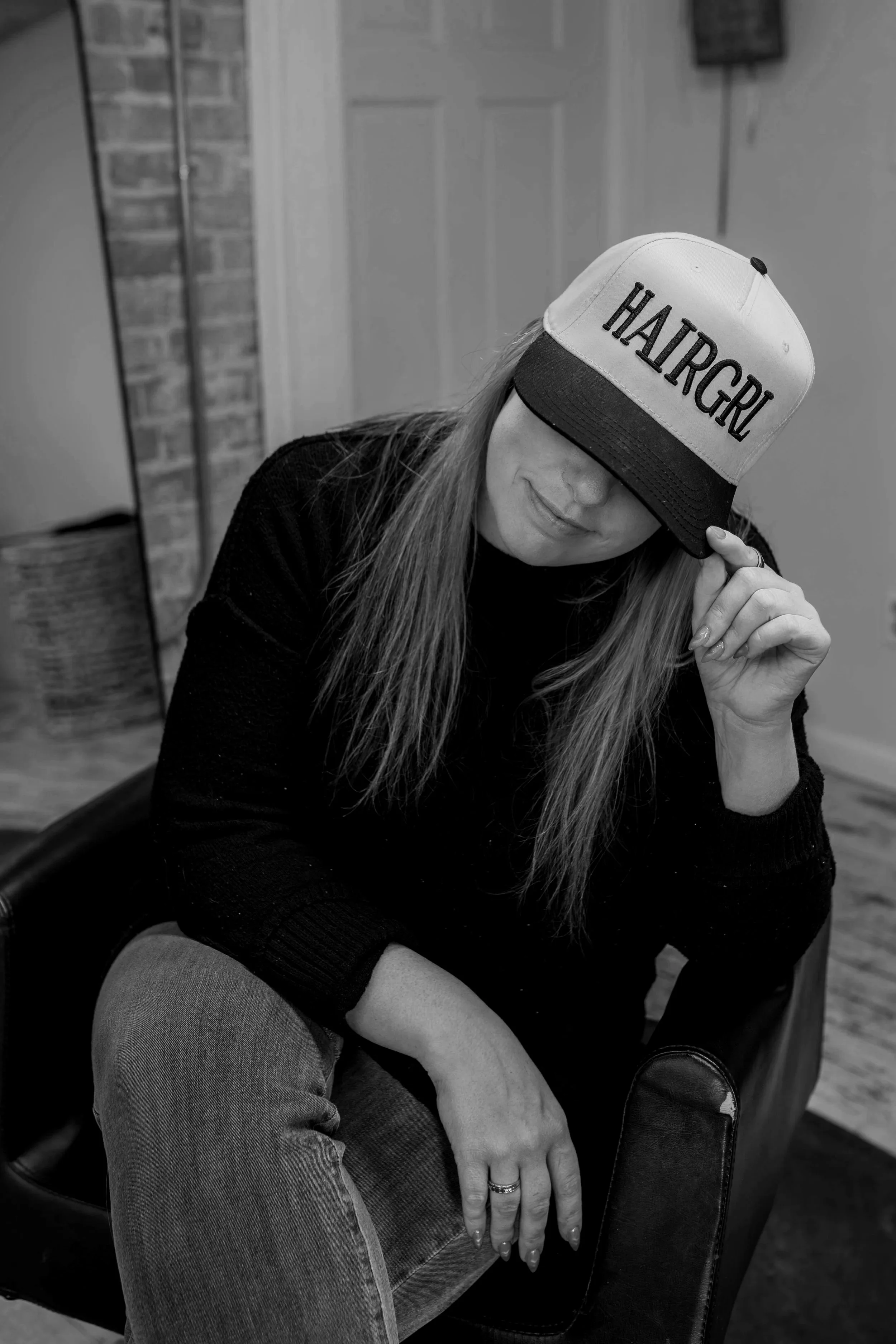 A woman wearing a cap that says 'HAIRGIRL' in large letters. She is sitting with one leg crossed over the other, her left hand resting on her knee. She has long hair and is dressed in a black top and ripped jeans. The photo is in black and white and appears to be taken indoors, with a closed door and a brick column in the background.