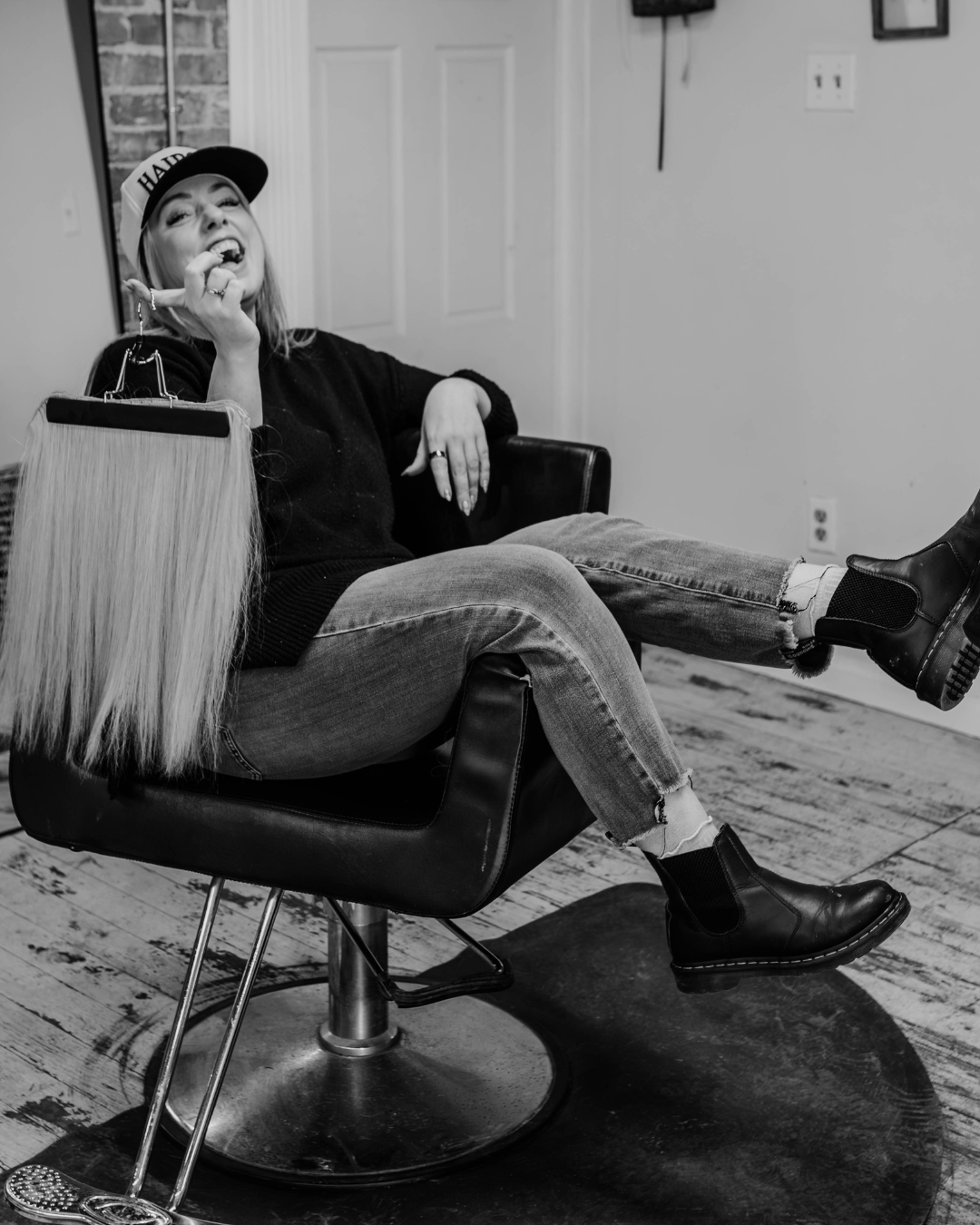 A woman with blonde hair, wearing a baseball cap, black sweater, ripped jeans, and black boots, sitting in a salon chair, smiling, with her feet up, and a hair extension comb on the floor in front of her.