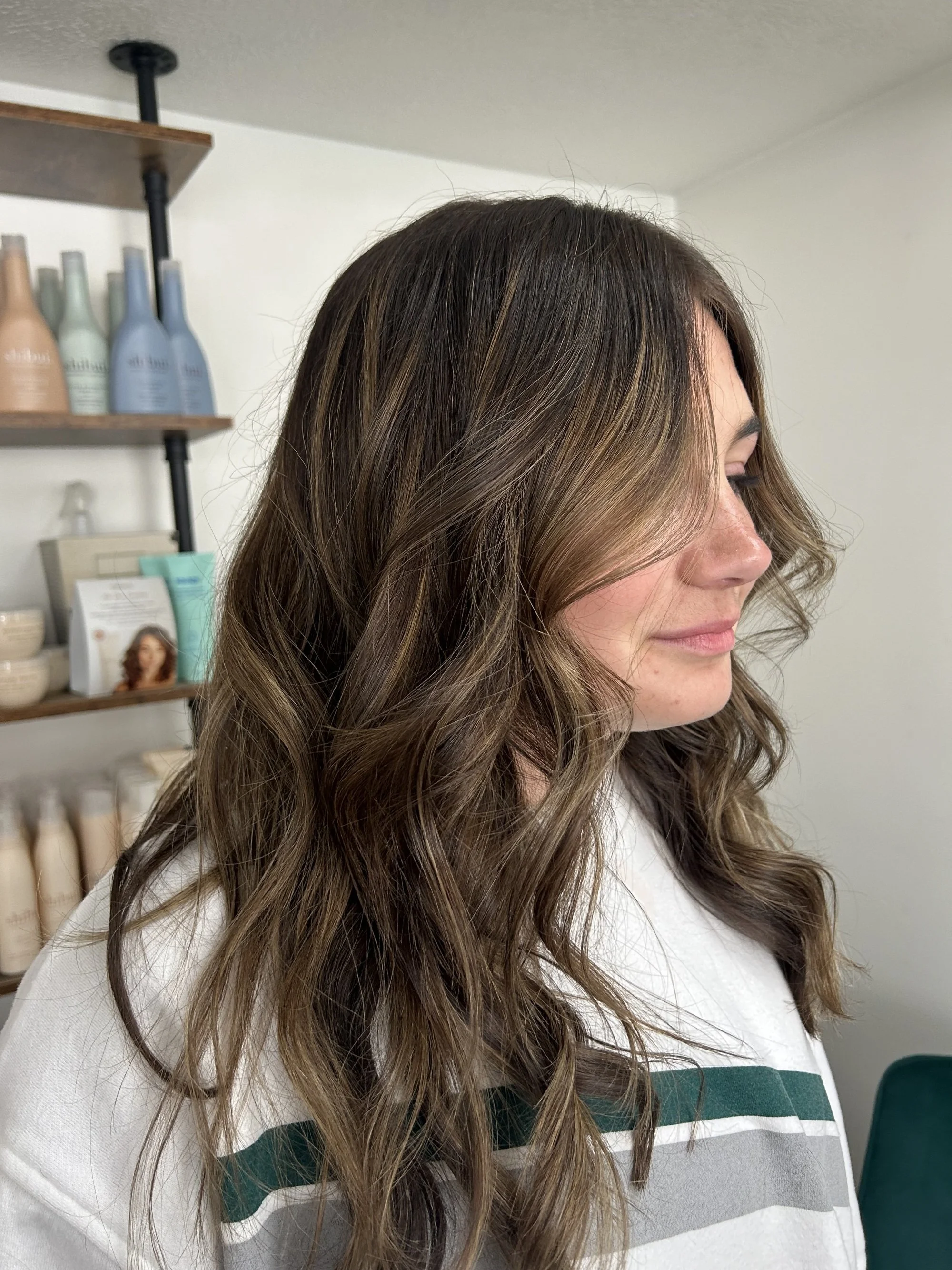 A woman with wavy, brown hair with blonde streaks, wearing a white shirt with green and gray stripes, is facing to the right.