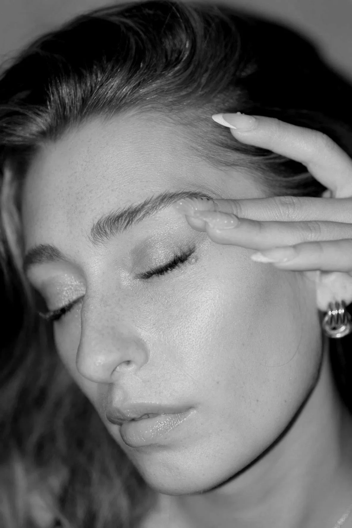 Close-up of a woman with closed eyes, her hand touching her forehead, showcasing her smooth skin, well-groomed eyebrows, and glossy lips in a black-and-white photo.