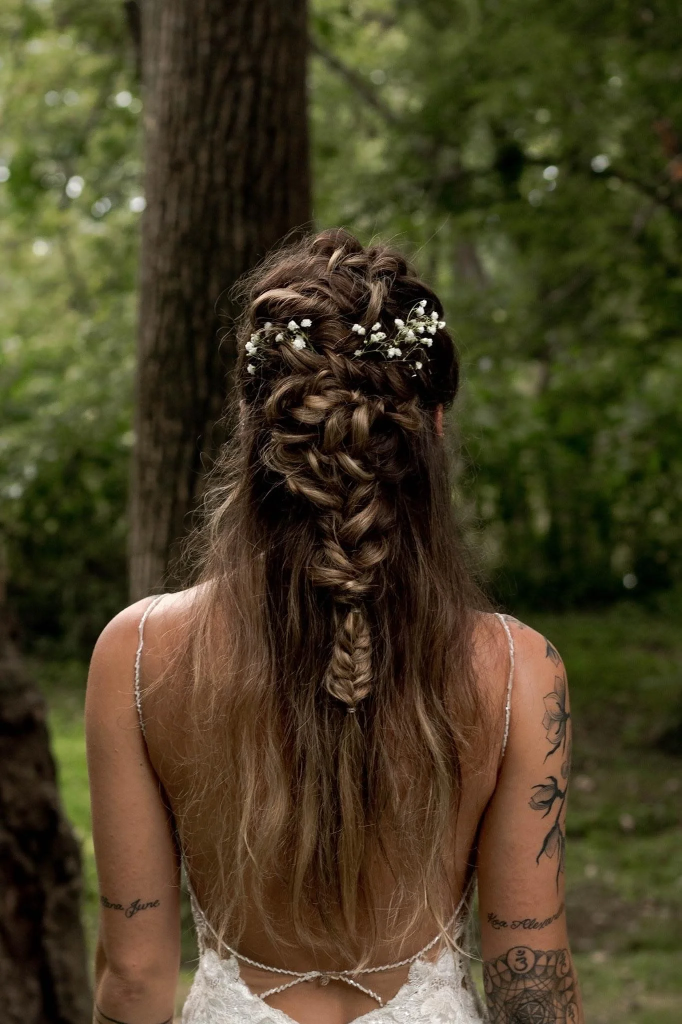 Back of a woman with long, wavy hair decorated with small white flowers, standing outdoors in a wooded area.