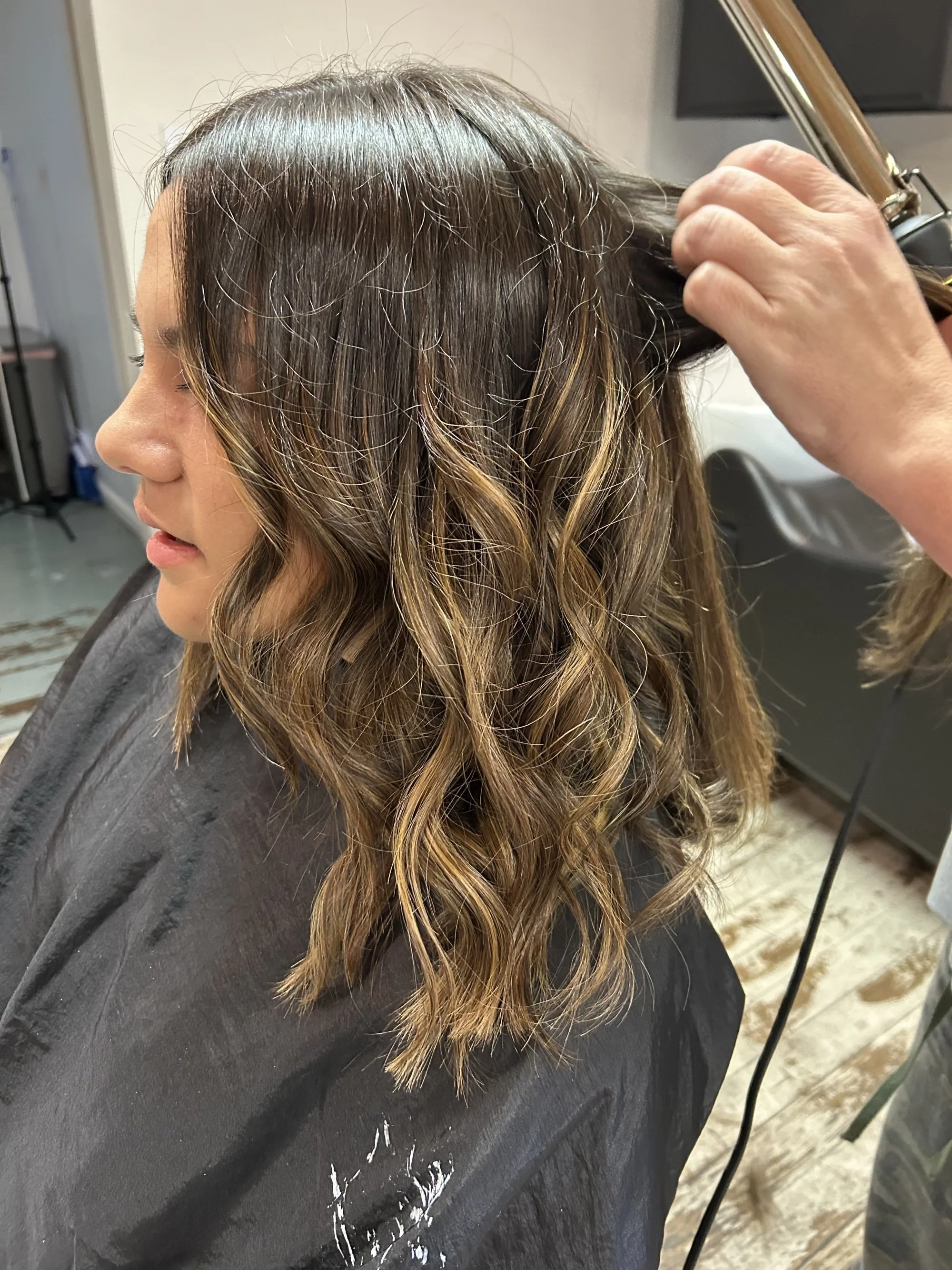 A woman at a salon getting her hair styled, with loose wavy highlights and a curling iron in her hand.