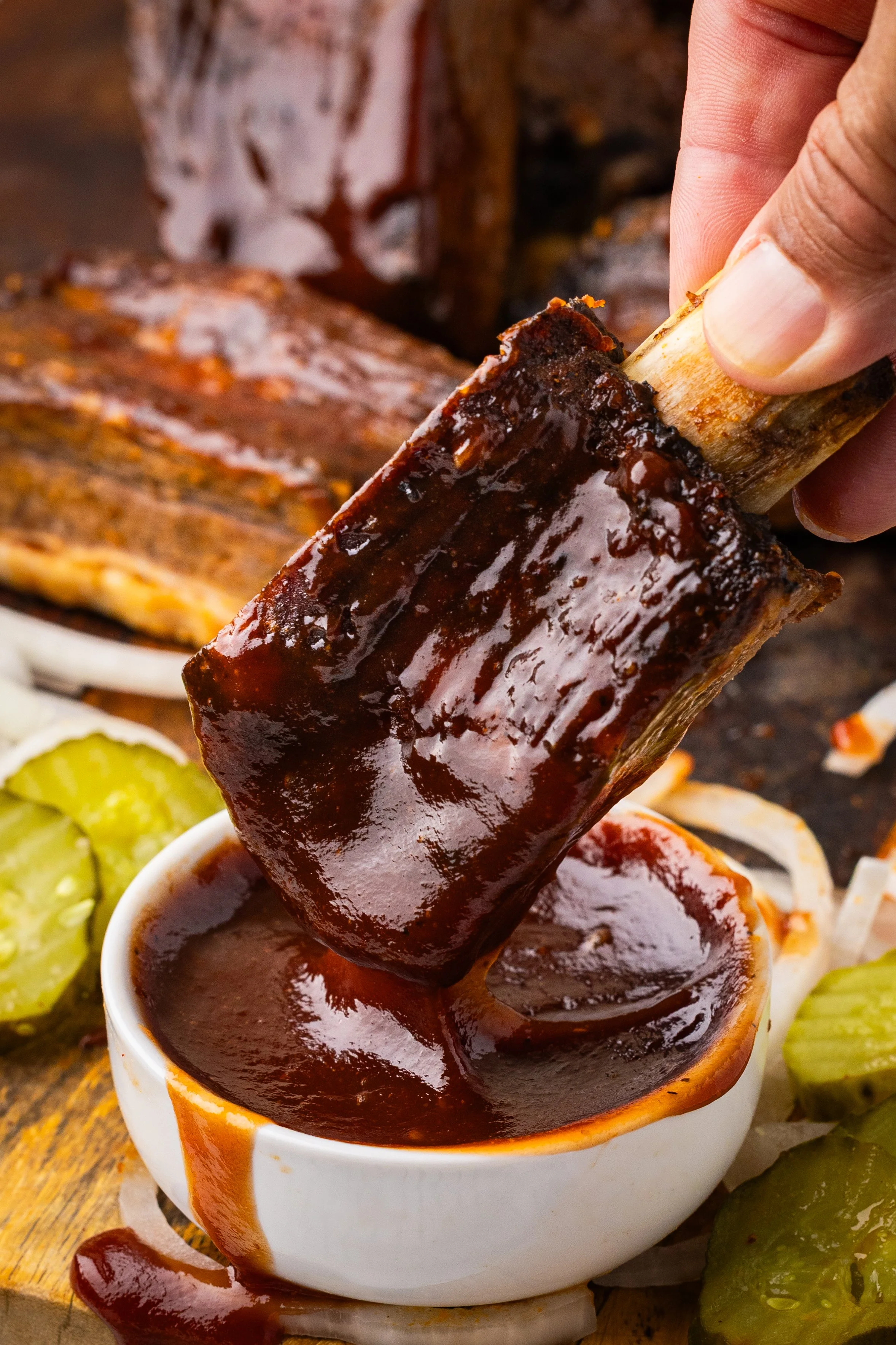 Beef Back rib being dipped into barbecue sauce with various side dishes including pickles.