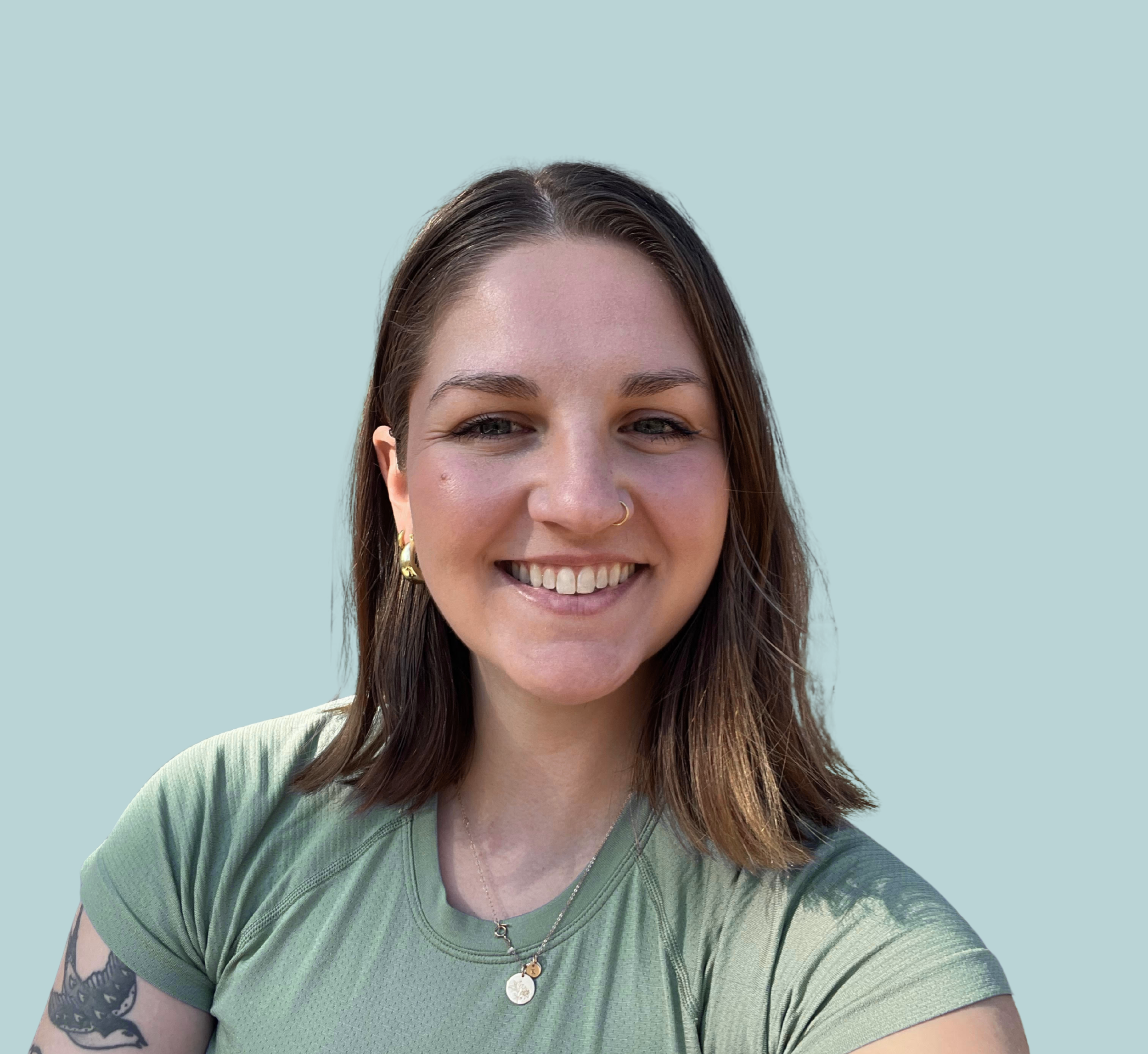 Close-up of a young woman with brown hair, smiling, wearing a green shirt, a necklace with two gold disks, gold hoop earrings, and a nose ring, against a light blue background.