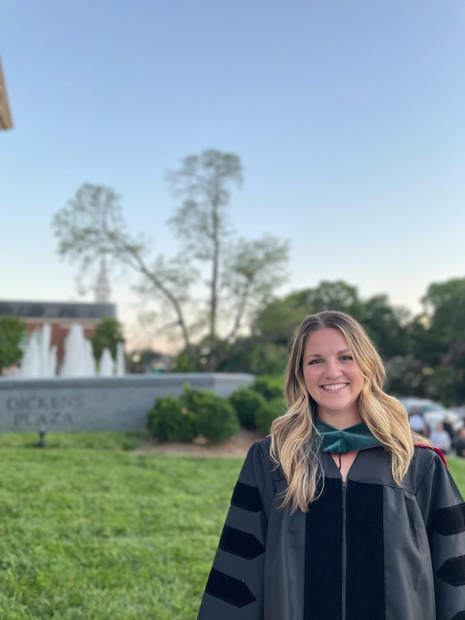 A young woman with blonde hair in a black graduation gown standing outdoors in front of a fountain and trees, smiling at the camera during daytime.